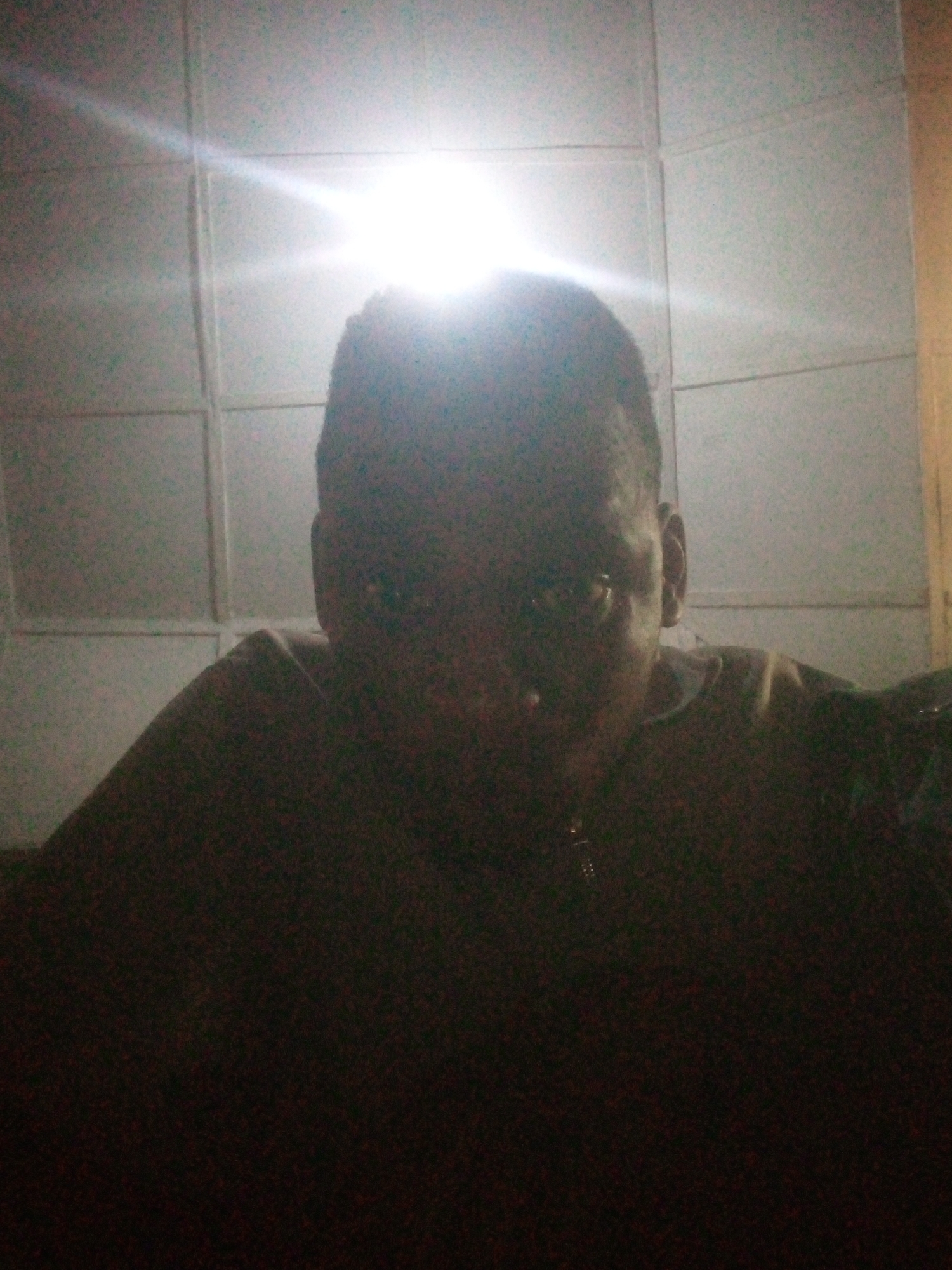 A dimly lit, indoor shot features a person with dark skin wearing glasses and a dark top. The person is positioned against a background of light-colored, rectangular tiles with seams. A strong light source above and behind the person casts a bright glow on the tiles and the top of the head, making it difficult to discern facial features. The location is Unknown, Unknown.