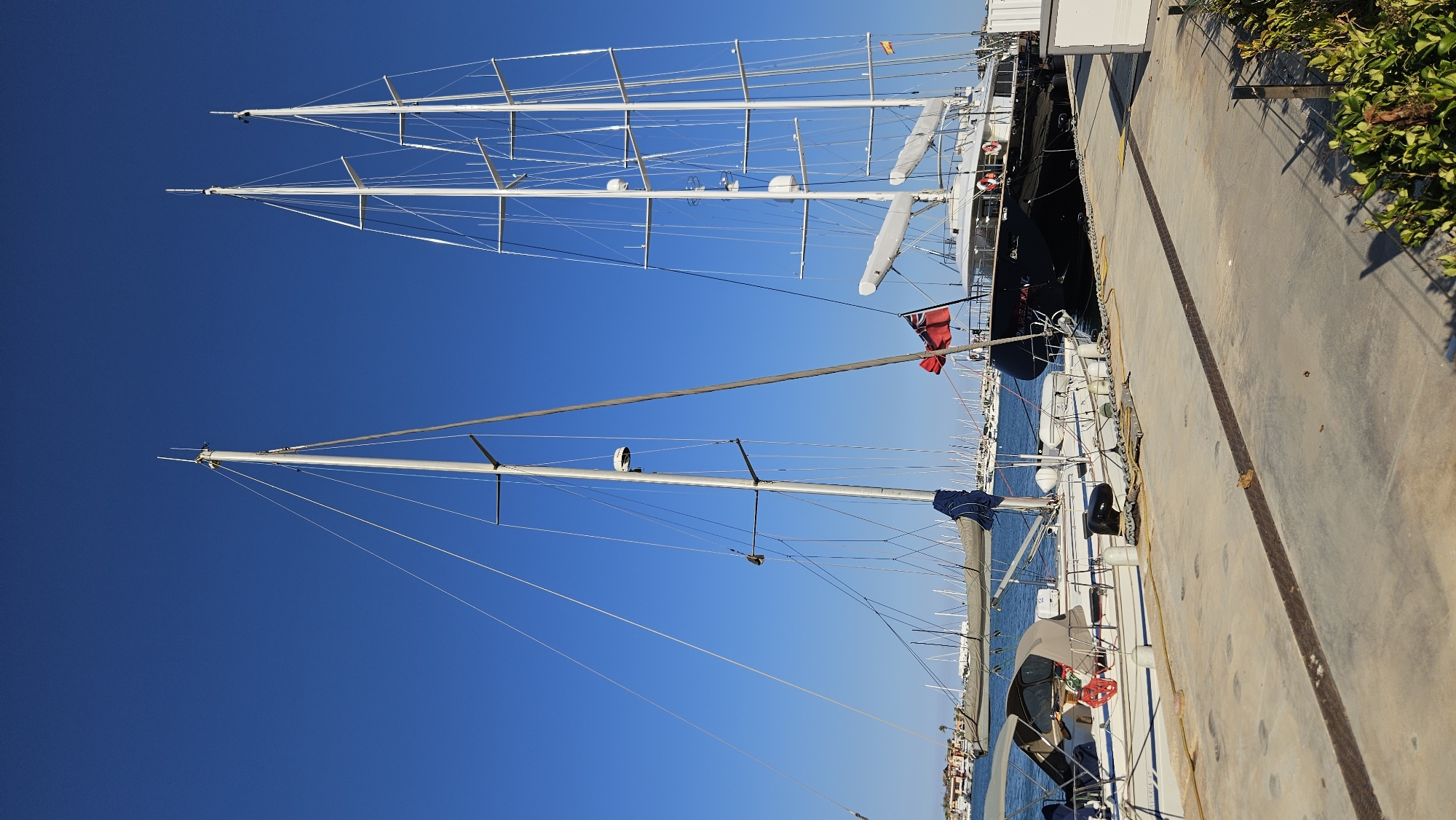 The image depicts a marina in Dénia, Spain, under a clear blue sky. In the foreground, a concrete pier is visible, with a metal strip embedded in its surface. A large, dark-hulled yacht with multiple masts dominates the right side of the frame. It flies a red Union Jack flag and appears to be moored. To the left of this yacht, a white sailboat is partially visible, also docked. In the background, numerous other sailboats and yachts fill the harbor, with buildings and palm trees lining the distant shore. The water is calm. The scene suggests a bright, sunny day at a bustling nautical location.