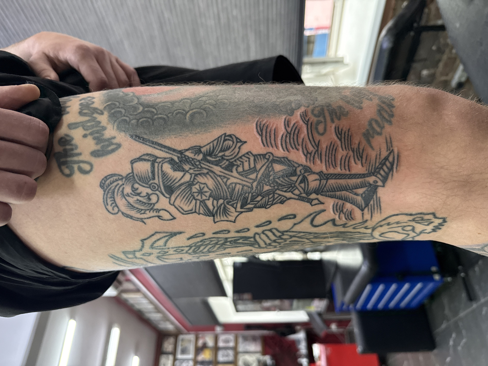 A close-up, slightly out-of-focus photograph shows a person's tattooed upper leg and knee. The main tattoo features a detailed image of a medieval knight lying horizontally with a sword in hand, depicted with heavy shading and line work. Above the knight, there's a cloud-like design. Above the clouds, there are parts of words "The bigger..." The leg is also tattooed with some abstract lines. The person's hand is visible in the upper left corner. The background is blurry and depicts interior elements like furniture. The location cannot be determined from the given photograph.