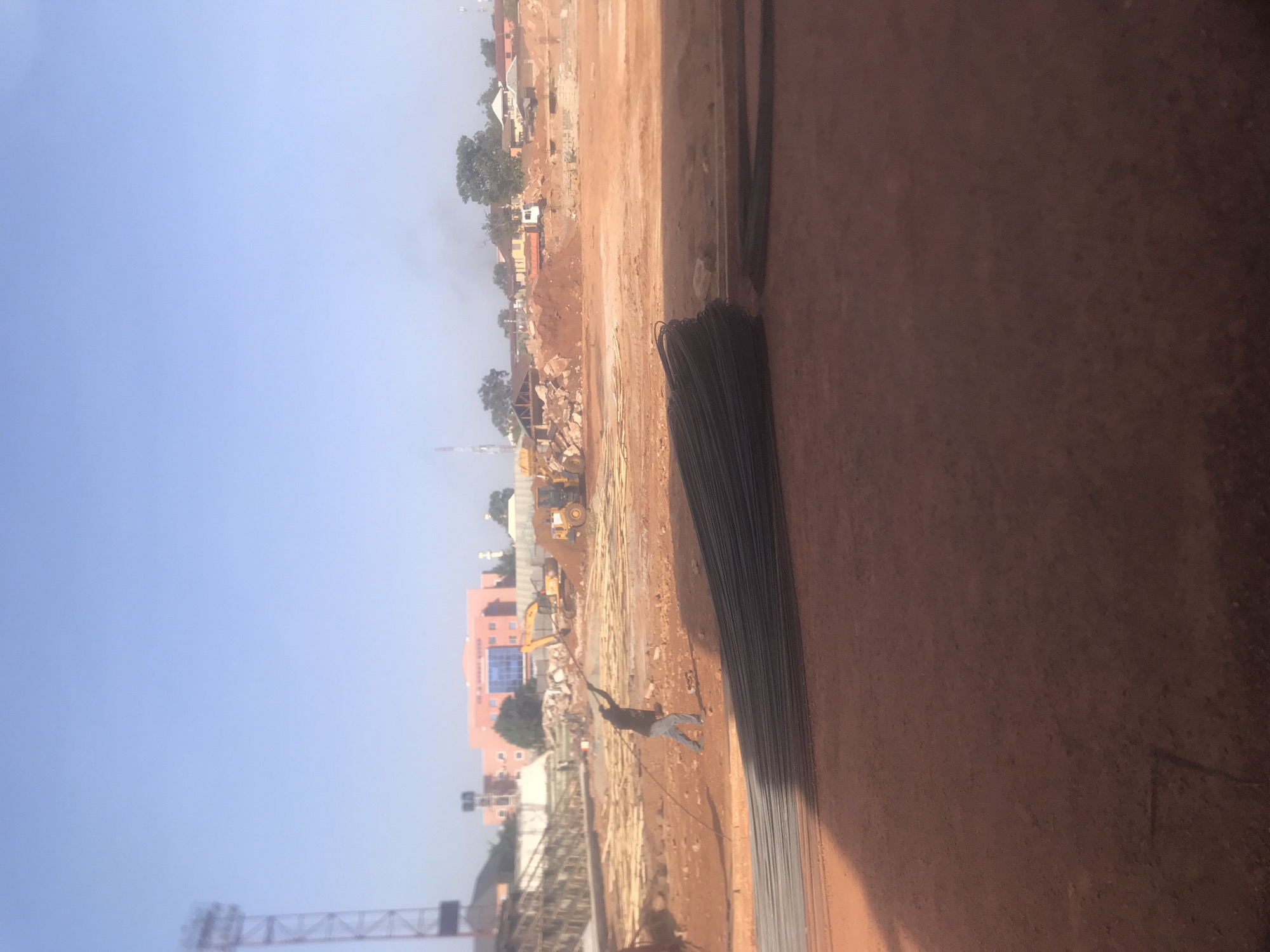 This image captures a wide view of a construction site under a clear, bright blue sky during the daytime. The ground consists primarily of reddish-brown earth, characteristic of the soil in Jos, Nigeria.
In the immediate foreground, to the right, there is a large, elevated section of reddish-brown soil or a wall. Adjacent to it, several bundles of steel reinforcement bars (rebar) are neatly stacked horizontally.
The middle ground is an expansive cleared area for construction. Piles of excavated dirt, rocks, and scattered wooden planks are visible across the site. Multiple pieces of heavy machinery, specifically two yellow excavators, are present, indicating ongoing work. One excavator is prominent in the mid-distance, and another is partially visible further back. A tall construction crane is also visible in the far-left background, suggesting a multi-story project.
A lone individual, dressed in a dark top and light trousers, stands in the center-left of the construction site. They are actively engaged with a long, dark cable or rope, appearing to be pulling or maneuvering it across the ground.
In the background, behind the construction activity, a cluster of buildings and trees lines the horizon. These include residential-style houses with reddish roofs and walls, and a larger, multi-story building with blue-tinted windows. A distinct plume of white smoke rises into the sky from the distant background, suggesting some form of industrial or burning activity. No visible text is present in the image. The scene portrays a phase of active development or land clearing.