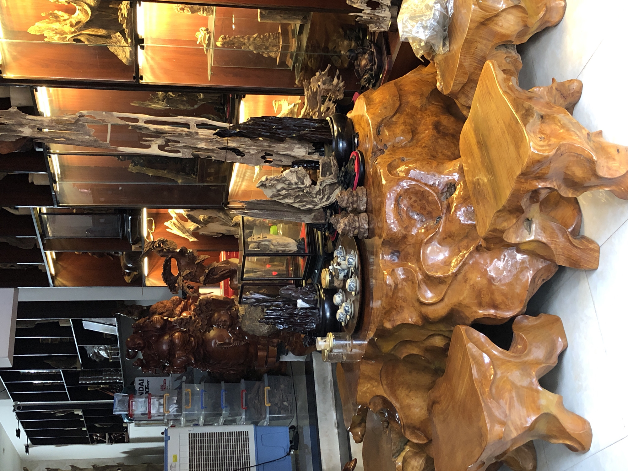 The image depicts the interior of what appears to be a shop or display area in Da Nang, Vietnam, specializing in wooden art pieces and furniture. In the foreground, a large, intricately carved wooden coffee table with organic, flowing lines is the central focus. Several wooden stools, also carved with a similar naturalistic style, are placed around the table. On the table, a small tea set, including cups and a teapot, is arranged on a dark tray.
Behind the table, a collection of wooden sculptures and decorative items are displayed. These include several abstract, twisted wood pieces and smaller carved figures. To the left, a large, dark wooden statue of a jovial figure, possibly a deity or a traditional character, stands prominently. Next to it, there are stacks of clear plastic storage containers.
The background features shelves filled with various wooden art pieces and decorative objects, some illuminated by warm lighting within glass display cases. The overall atmosphere suggests a curated collection of handcrafted wooden art, likely for sale. The environment appears to be indoors, with tiled flooring visible in the foreground. There are no people visible in the image.