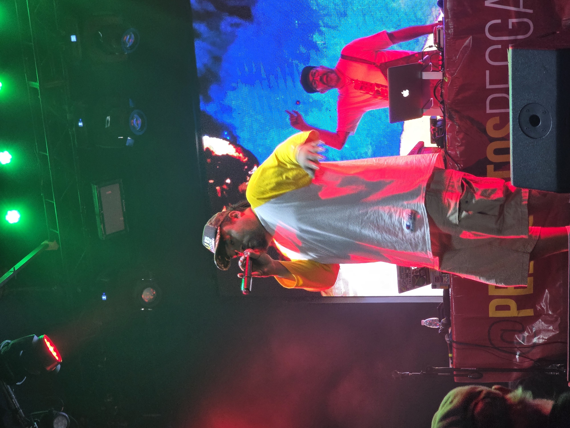 This image depicts a live music performance on a stage within a dark indoor venue in Buenos Aires, Argentina. The primary subject is a male performer in the foreground, characterized by a dark baseball cap, a white t-shirt with yellow long sleeves, and light-colored shorts. He has a beard and is actively holding a black microphone, which displays red and green illumination, to his mouth as if singing or speaking. Behind him, to the right, a second male individual, identified as a DJ, wears a dark flat cap, glasses, a light red shirt, and dark suspenders. He stands behind a table laden with electronic equipment, including a silver Apple MacBook on a stand, other audio devices (one partially labeled "ALLEN & HEATH," another "DUB TEKNIK"), and cables, while gesturing with his raised right index finger. The table itself is draped in red fabric featuring yellow text that partially reads "PEñATOSDEREGGAE." A small, clear plastic water bottle is also present on the table. The background consists of a large LED screen displaying abstract patterns of blue, white, and hints of red, resembling illuminated smoke or fire. The stage is overhead lit by multiple red and green stage lights mounted on a truss system, with visible atmospheric haze. A black stage monitor speaker is positioned in the lower foreground. A partial view of an audience member wearing a green hat is visible in the bottom left corner.
