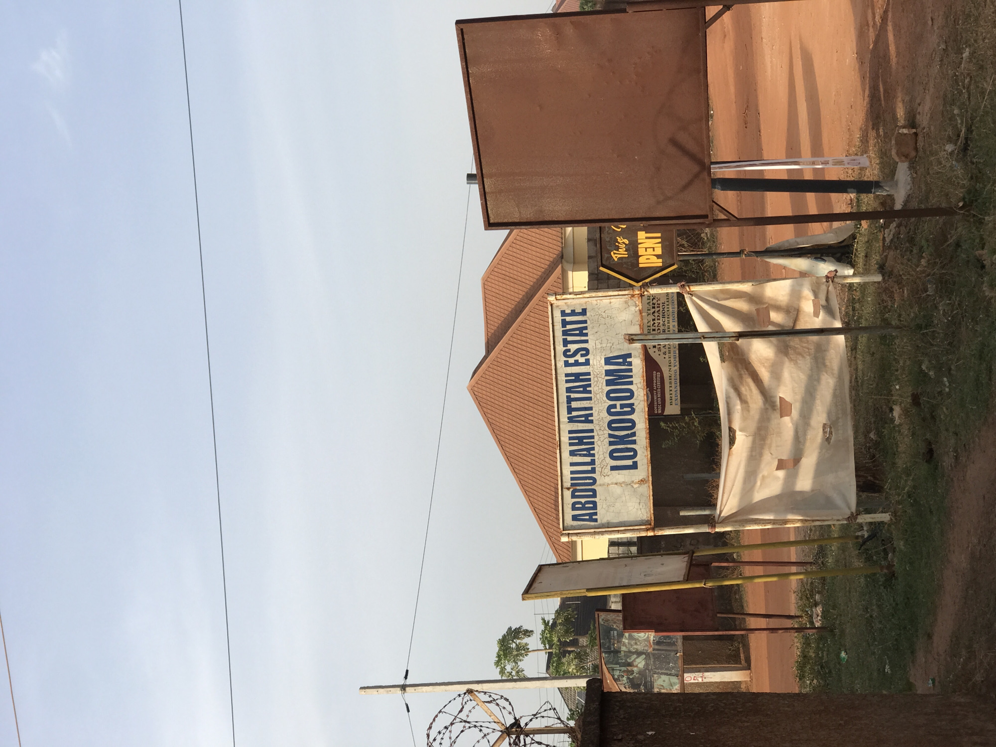 The image depicts a bright daytime street scene, likely a roadside in a developing area, characterized by several signs and a building under a clear sky. In the center, a building with a reddish-brown tiled roof is visible.
Along the roadside, multiple signs are present. A prominent, weathered sign in the mid-ground, featuring blue text on a white background, reads "ABDULLAHI ATTAH ESTATE LOKOGOMA." To its right, another sign, partially obscured, contains text suggesting an educational institution, including phrases like "GOVERNMENT APPROVED," "WAEC AND NECO CERTIFIED," "BRITISH/NIGERIA," and fragments such as "EXPANDING YOUR C," "RLY YEAR" (Early Year), "IMARY" (Primary), "ONDARY" (Secondary), "R-SCHOOL," "IRRICULAR" (Curricular), and "D'S HORIZONS." Further right, a small, hexagonal yellow and black sign points with text "This & IPENT," possibly indicating an event or direction. A large, plain brown billboard stands behind these signs.
The ground is a reddish-brown dirt road with patches of dry grass. A tattered white banner or cloth is suspended between two thin metal poles on the right side of the "Abdullahi Attah Estate" sign. On the far left, a concrete utility pole wrapped with barbed wire is visible, with a single power line stretching across the light blue sky. The scene is devoid of people. The clear sky and bright illumination suggest it is mid-day. The provided context indicates the location is Abuja, Nigeria.