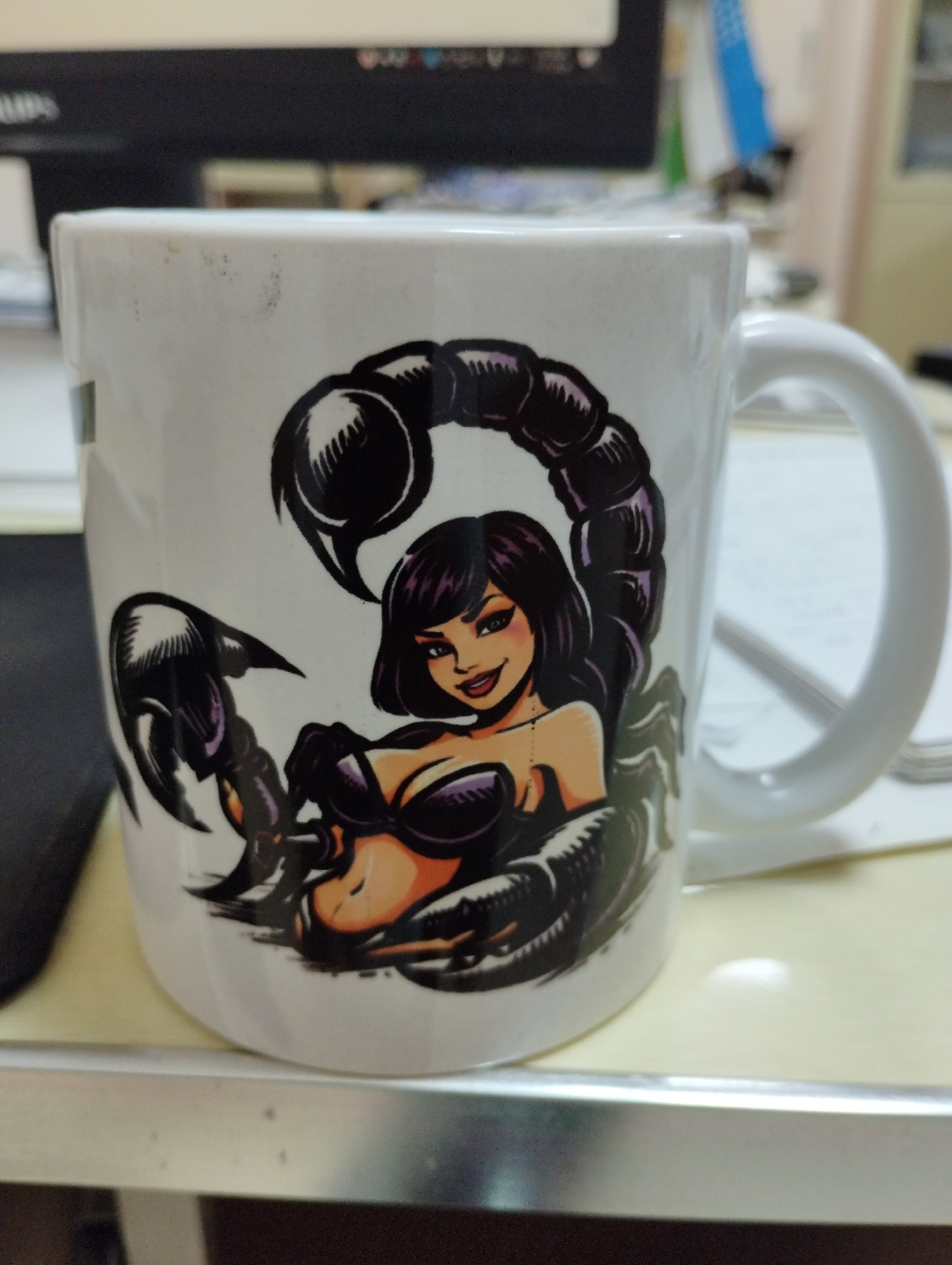 A white ceramic mug with a graphic design of a scorpion-woman is placed on a light-colored desk. The graphic depicts a woman with dark hair and a black scorpion tail curling around her. She is wearing a black top and has a playful expression. The mug has a white handle. In the background, a computer monitor is visible, along with some papers and office supplies. The overall setting appears to be an office environment. The image does not contain any information about the city of Urgench, Uzbekistan.