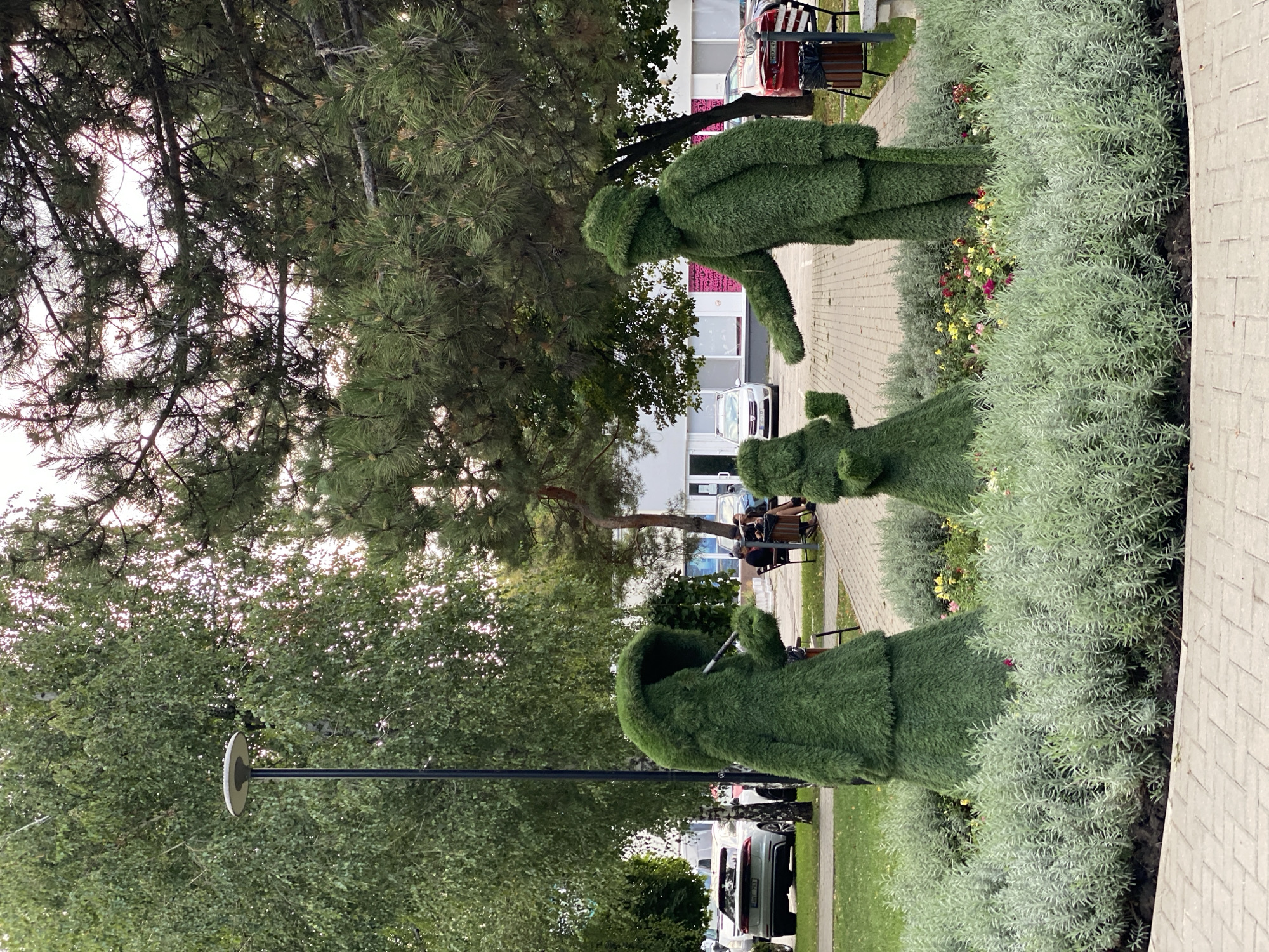 The image shows a streetscape in Chișinău, Moldova, with a focus on three topiary figures resembling people. The figures are made of green foliage and are arranged in a garden bed. The leftmost figure is tall and wears a hood, appearing to hold a thin stick or pipe. In the center is a smaller figure. To the right, a taller figure in a hat gestures outwards. Behind the topiaries, a paved walkway is visible, with park benches and people seated on them. Further back, several cars are parked, including a white Audi and a red car. Buildings with windows and a street lamp are also present in the background. The scene is set amidst lush green trees and vegetation.
