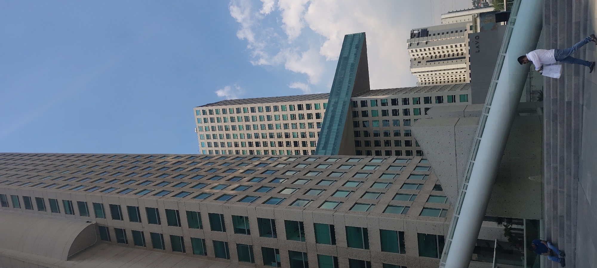 The image shows a collection of modern architectural structures in Mexico City, Mexico. The buildings feature grid-like facades with numerous windows. A prominent element is a vertical glass structure connecting two buildings. A pedestrian bridge is visible on the right side of the frame. A man in a white shirt and jeans is walking on the bridge, carrying a white bag. Another individual is seated on the steps of the bridge. The sky is partly cloudy. "LAVO" is visible on a building near the bridge.