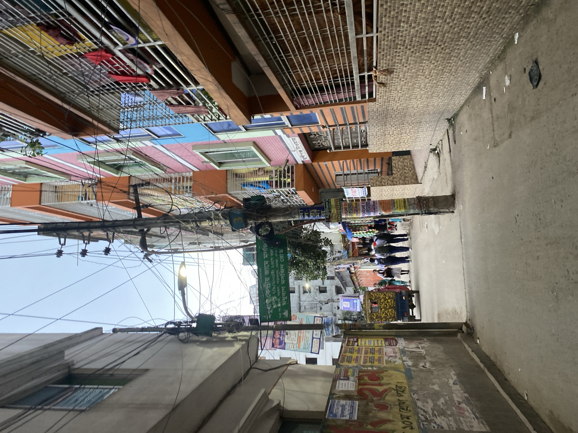The image depicts a street scene in Dhaka, Bangladesh, as indicated by the street sign "Madhubagh Masjid Road." The narrow street is bordered by buildings, with one side featuring a brick structure and the other a series of colorful, multi-story buildings with balconies and laundry hanging out to dry. A power pole laden with wires dominates the center of the frame, obscuring the sky. Below, a group of people walks along the street, past an auto rickshaw. The street wall shows evidence of posters and advertisements.