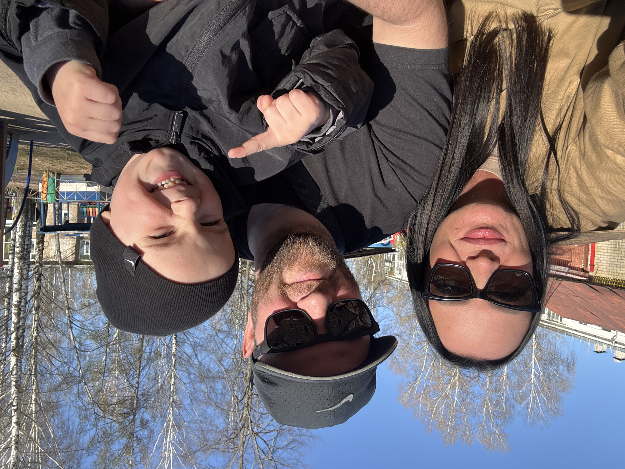 A selfie taken from a low angle in an outdoor setting shows a family of three. A young boy in a black beanie and jacket is positioned on the left. A bearded man wearing sunglasses and a black cap is in the center, and a woman with long dark hair and sunglasses is on the right. All three appear to be smiling. The background consists of leafless trees, blue sky, and buildings. The scene is outdoors, likely in a park or public area. The location is identified as the city of Chaikovsky, Russia.