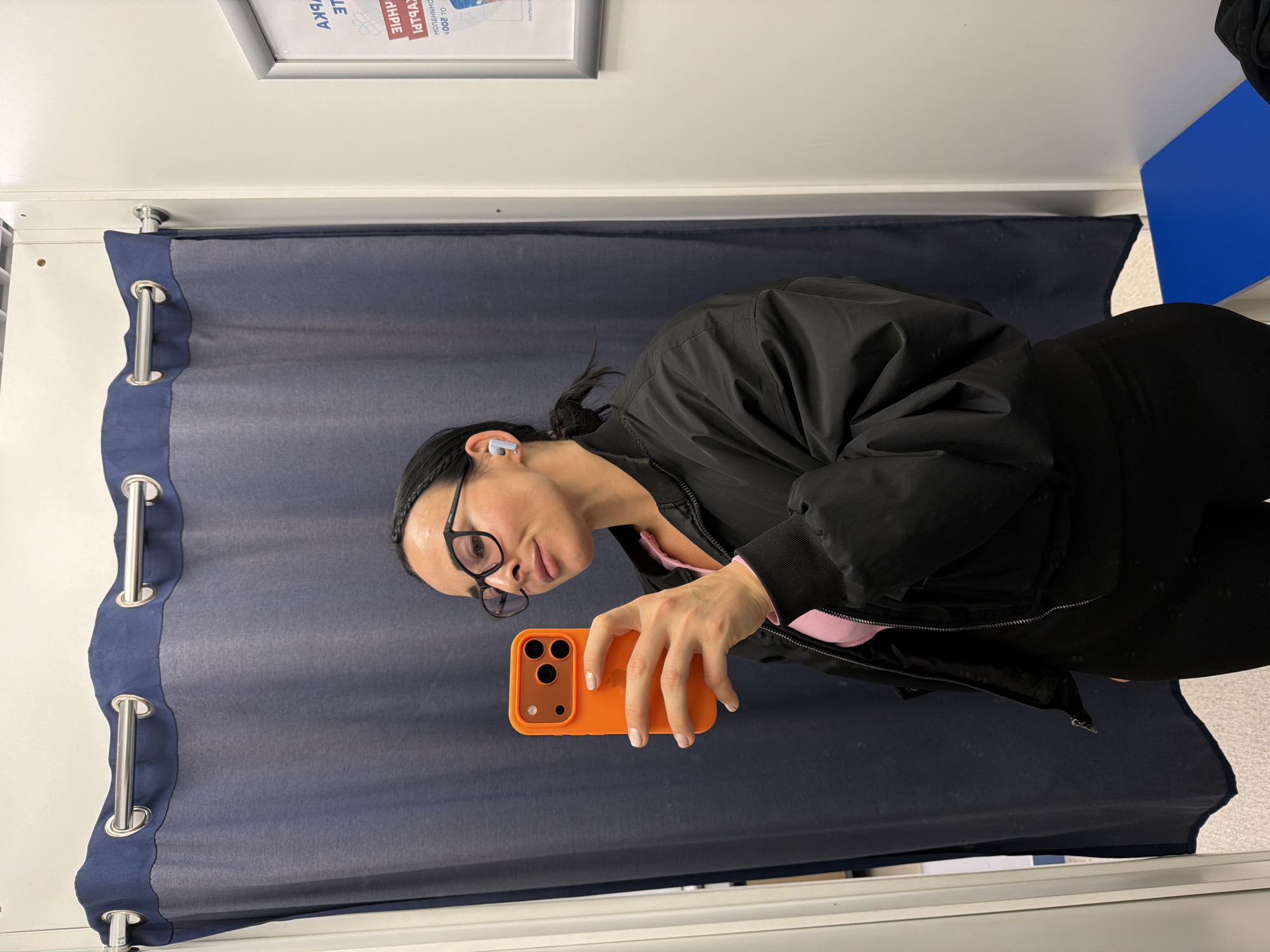 A woman with dark hair pulled back, wearing glasses, an earbud, a pink shirt, and a black jacket, is holding an orange iPhone in what appears to be a changing room. The room has a dark blue curtain with silver grommets along the top edge. The walls are white and a blue shelf is visible in the background. A framed poster hangs on the wall above the curtain rod, with Russian text including the words "Санкт-Петербург" (Saint Petersburg). Therefore, it is safe to assume the photograph was taken in Saint Petersburg, Russia.