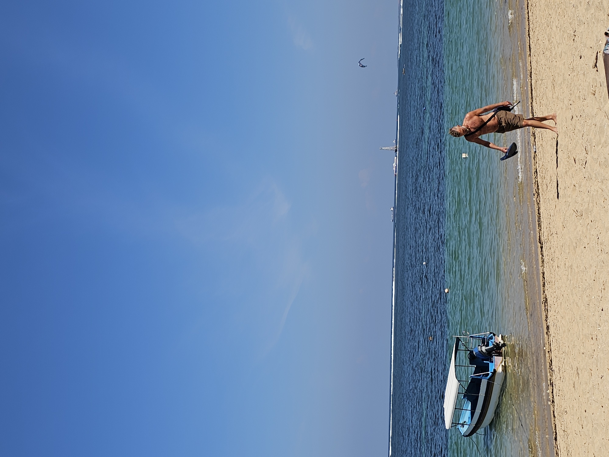A shirtless man walks along a sandy beach with a blue and white boat moored in the shallow turquoise water to his left. The man is wearing brown shorts and carrying a dark hat and a dark bag over his shoulder. In the background, the ocean extends to the horizon, with a white wave line visible. Several boats and a tall structure are visible on the water further out. The sky is a clear, bright blue with a few wispy clouds. The scene appears to be Sanur Beach in Denpasar, Indonesia, known for its calm waters and sunrise views.