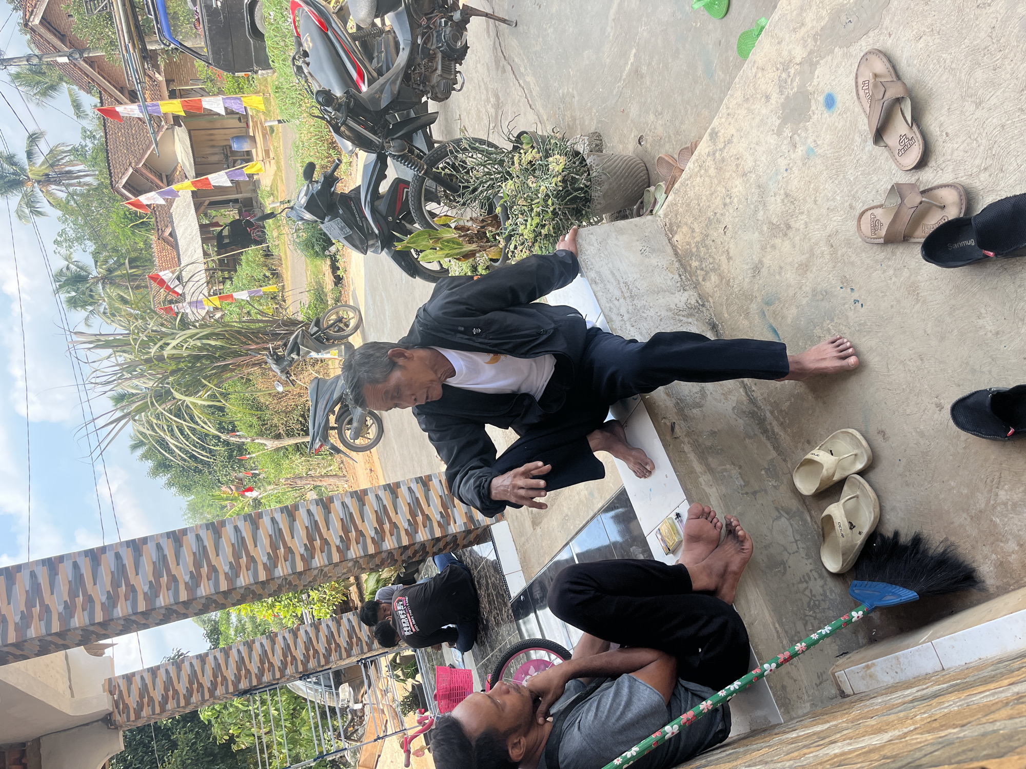 Two men are sitting on the steps of a house in Karang Sari, Indonesia. The man on the left is wearing a grey t-shirt and black pants, and is looking towards the man on the right. The man on the right is wearing a black jacket over a white t-shirt and black pants, and is speaking to the man on the left. Behind the two men, another two men are sitting on the floor. In the background, there are several motorcycles parked, houses with red tile roofs, and palm trees. The weather is sunny with some clouds. The steps of the house are decorated with colorful tiles. There is a broom leaning against the wall.