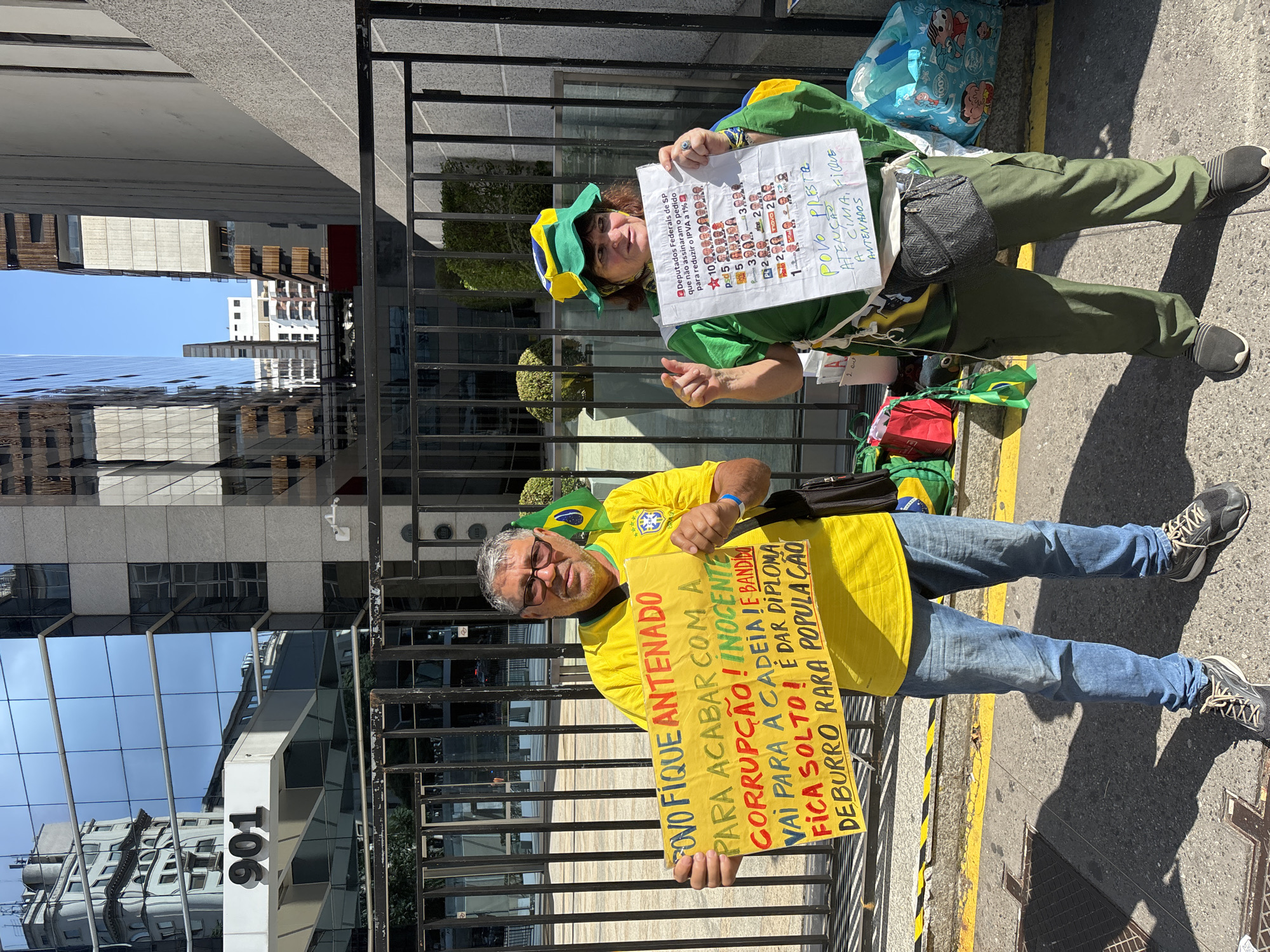 The image shows two individuals, a man and a woman, participating in a street protest or demonstration on a sunny day in an urban environment, identified as São Paulo, Brazil. They are standing on a paved sidewalk next to a dark metal fence, with modern glass buildings reflecting the clear blue sky in the background, one of which displays the number "901".
The man, appearing middle-aged with grey hair and glasses, is wearing a yellow t-shirt with a Brazilian flag logo, blue jeans, and dark athletic shoes. He holds a rectangular yellow sign that reads: "POVO FIQUE ANTENADO PARA ACABAR COM A CORRUPÇÃO! INOCENTE VAI PARA A CADEIA E BANDIDO FICA SOLTO! É DAR DIPLOMA DE BURRO PARA POPULAÇÃO" (People, stay alert to end corruption! Innocent goes to jail and bandit goes free! It's giving a diploma of donkey to the population). He also wears a black shoulder bag.
The woman, also appearing middle-aged, is dressed in a green t-shirt, green cargo pants, and dark athletic shoes. She wears a hat designed with the Brazilian flag colors (green, yellow, blue) and a green sash or vest. She holds a white sign with two main texts. The top section reads: "Deputados Federais de SP que não assinaram o pedido para reduzir o IPVA a 1%" (Federal Deputies from SP who did not sign the request to reduce the IPVA (Vehicle Property Tax) to 1%), followed by a list of political parties or figures with numerical indications. The bottom part urges: "POVO PRESTE ATENÇÃO! FIQUE ANTENADOS" (People, pay attention! Stay alert). She also has a small patterned waist bag and a light blue tote bag featuring Monica's Gang characters.
Both individuals' attire, incorporating the Brazilian national colors, signifies their participation in a politically charged event, likely expressing anti-corruption sentiments and advocating for specific legislative changes.