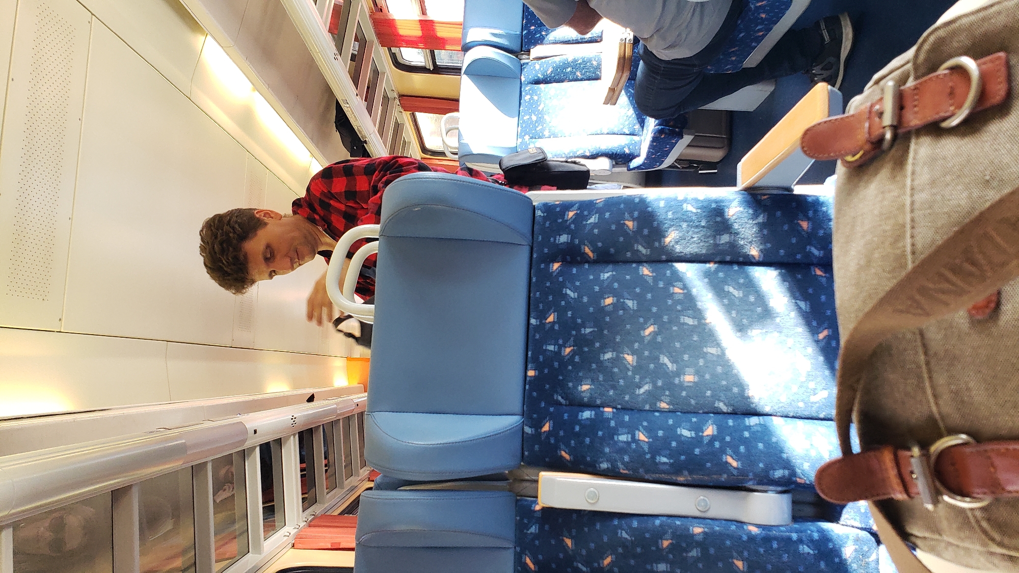 A man with curly brown hair wearing a red and black checkered shirt is standing on a train and adjusting a black bag. He is in the foreground, with the seats of the train in front of him and the ceiling of the train above. The seats are blue with a blue and orange pattern. The train is well-lit with natural light coming through the windows. The man appears to be in the process of stowing or retrieving an item from his bag. The scene is likely within a train carriage, which could be travelling through or stopping at the town of Štrba, Slovakia, as the image metadata suggests.