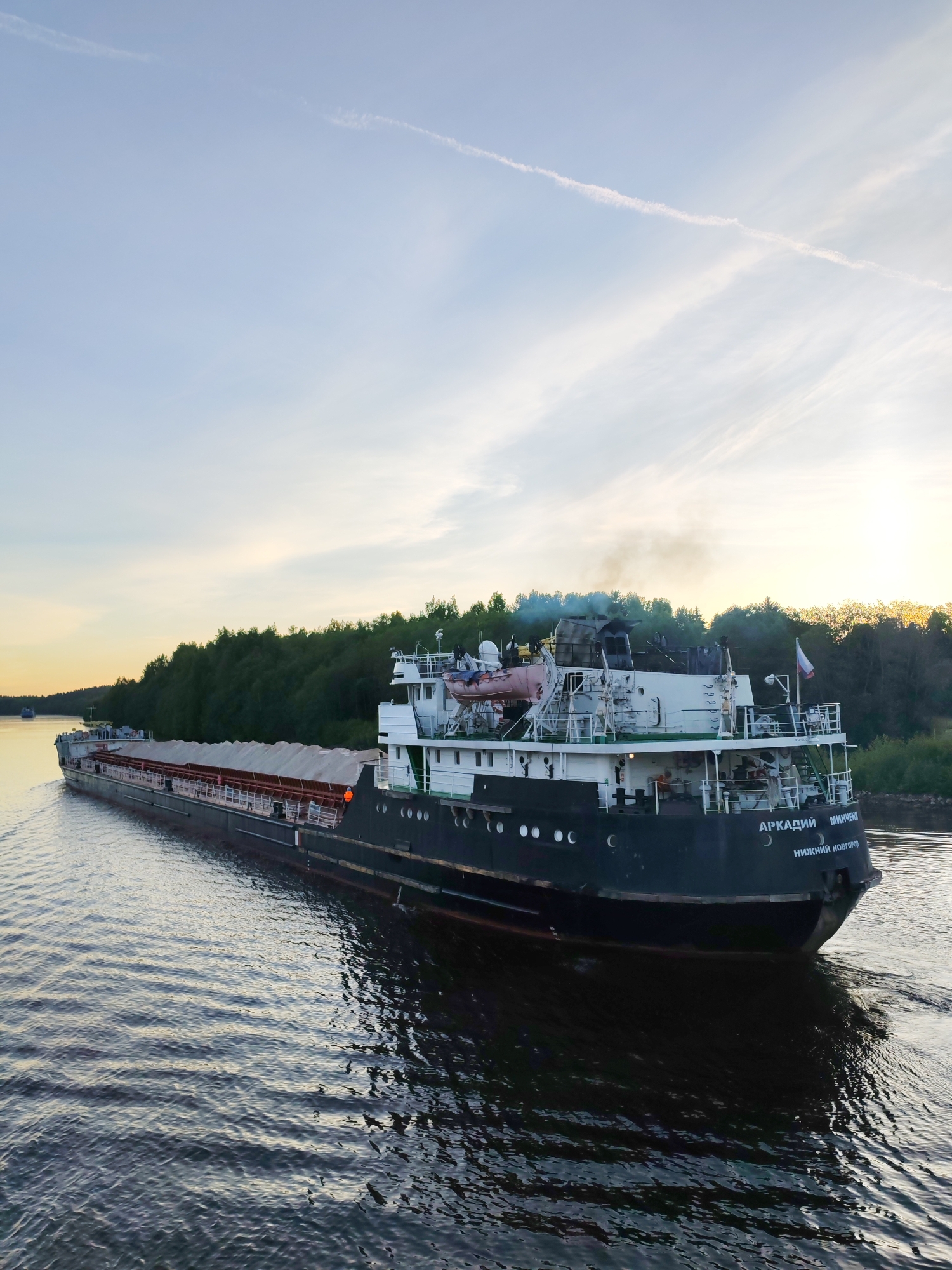 A long cargo ship, identified as "АРКАДИЙ МИНЧЕН" (ARKADY MINCHEN) from its home port of "НИЖНИЙ НОВГОРОД" (NIZHNIY NOVGOROD), navigates a wide river or canal at Volokov Most, Russia. The vessel has a dark hull and a white superstructure at its stern, from which light smoke or exhaust emanates. A Russian flag is visible near the stern. The ship appears to be loaded with bulk cargo, covered by white tarpaulins or covers across its numerous holds.
The scene is captured during either late afternoon or early evening, as indicated by the warm, low sunlight casting a golden glow on the distant treeline and reflected faintly on the water. The sky is a soft blue with subtle wisps of clouds and a faint contrail. The ship moves from the upper left towards the lower right of the frame, leaving a clear wake behind it. The water is calm, reflecting the sky and the ship's dark hull. A dense, dark green forest lines the right bank of the waterway. Another, smaller boat is barely visible in the distance further along the river. The overall atmosphere is peaceful and industrious.