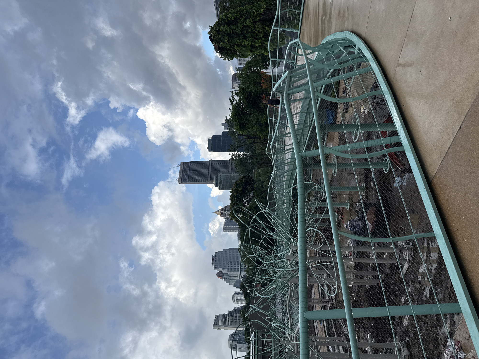 A slightly tilted, eye-level outdoor shot captures a view of Bangkok, Thailand. The foreground features a section of a pedestrian walkway or overpass with a green-painted metal safety fence adorned with decorative circular and linear elements.
Beyond the fence, a construction site is visible. The city skyline is comprised of modern high-rise buildings of varying heights and architectural styles. Trees with dense green foliage are interspersed among the buildings.
The sky is a mix of blue and cloud cover. White, puffy clouds are scattered throughout, with darker, more ominous-looking clouds concentrated in sections of the sky.