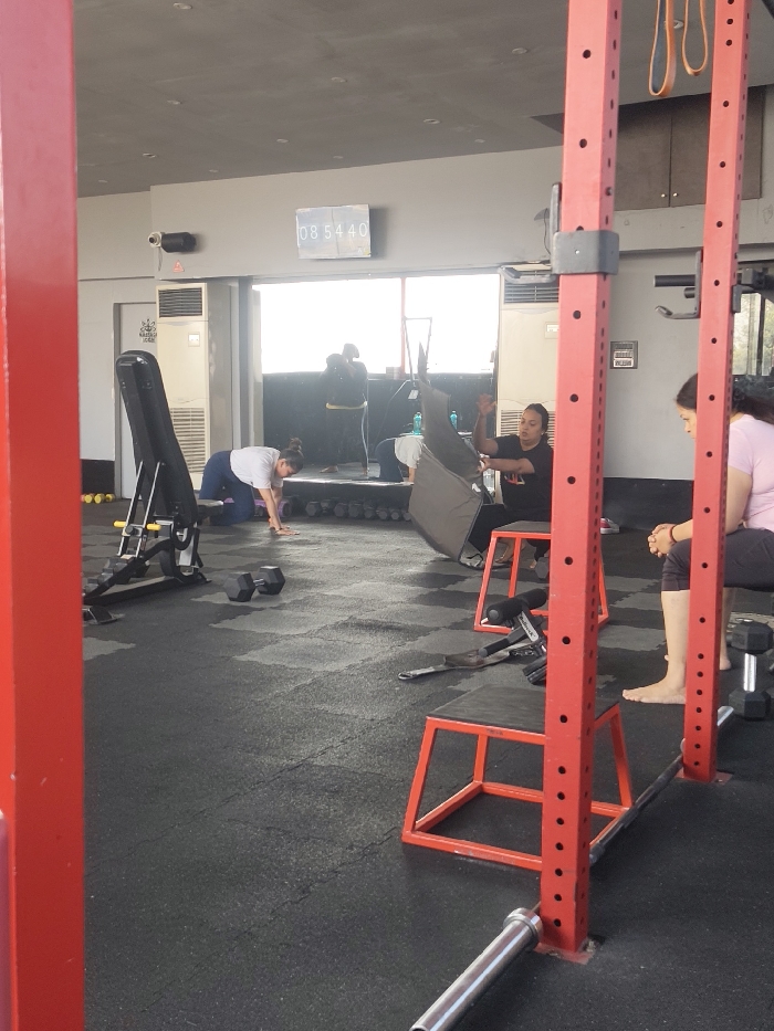 In a gym setting, multiple individuals are engaged in various activities. One person is kneeling on the ground, while another is seated on a stool holding an object. In the background, other individuals are visible near a window. The gym contains weightlifting equipment, including a barbell and squat rack. A digital clock displays "08 54 40" on the wall.