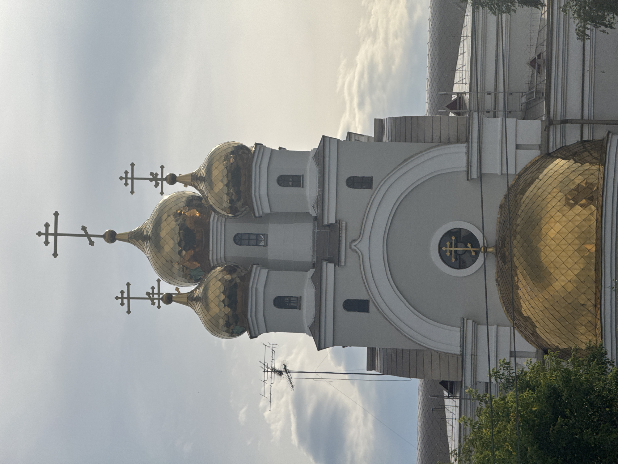 The image shows the Trinity Cathedral in Yekaterinburg, Russia, on a cloudy day. The cathedral has several golden domes, each topped with a cross. The main dome is large and prominent, while smaller domes are visible on the upper levels of the structure. The building is light grey with white architectural details. In the foreground, trees with green foliage are visible. An antenna is mounted on a pole to the left of the cathedral, extending into the sky. The sky is overcast with scattered clouds.