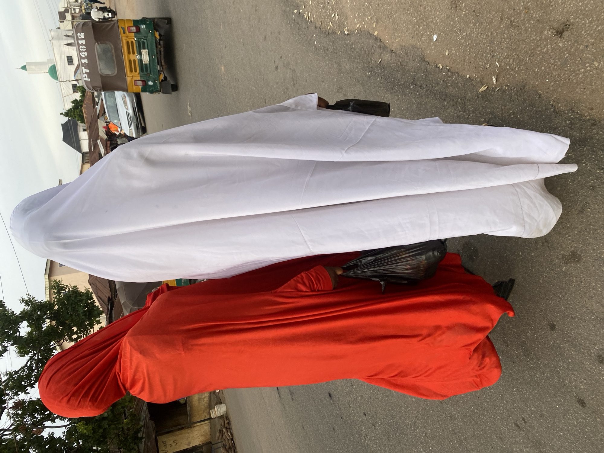 Two individuals, one wearing a bright red garment and the other a white garment, are walking on a paved road in Jos, Nigeria. The person in red is on the left, holding a black plastic bag in their right hand. The person in white is on the right. Both are facing away from the camera. In the background, there are buildings, including one with a green dome and a white minaret, indicative of a mosque. A yellow and green auto-rickshaw with license plate "PT-14812" is driving on the road. Other vehicles and a few people are also visible in the distance. The sky is overcast. The road appears to be a commercial area with some stalls and structures lining the sides.