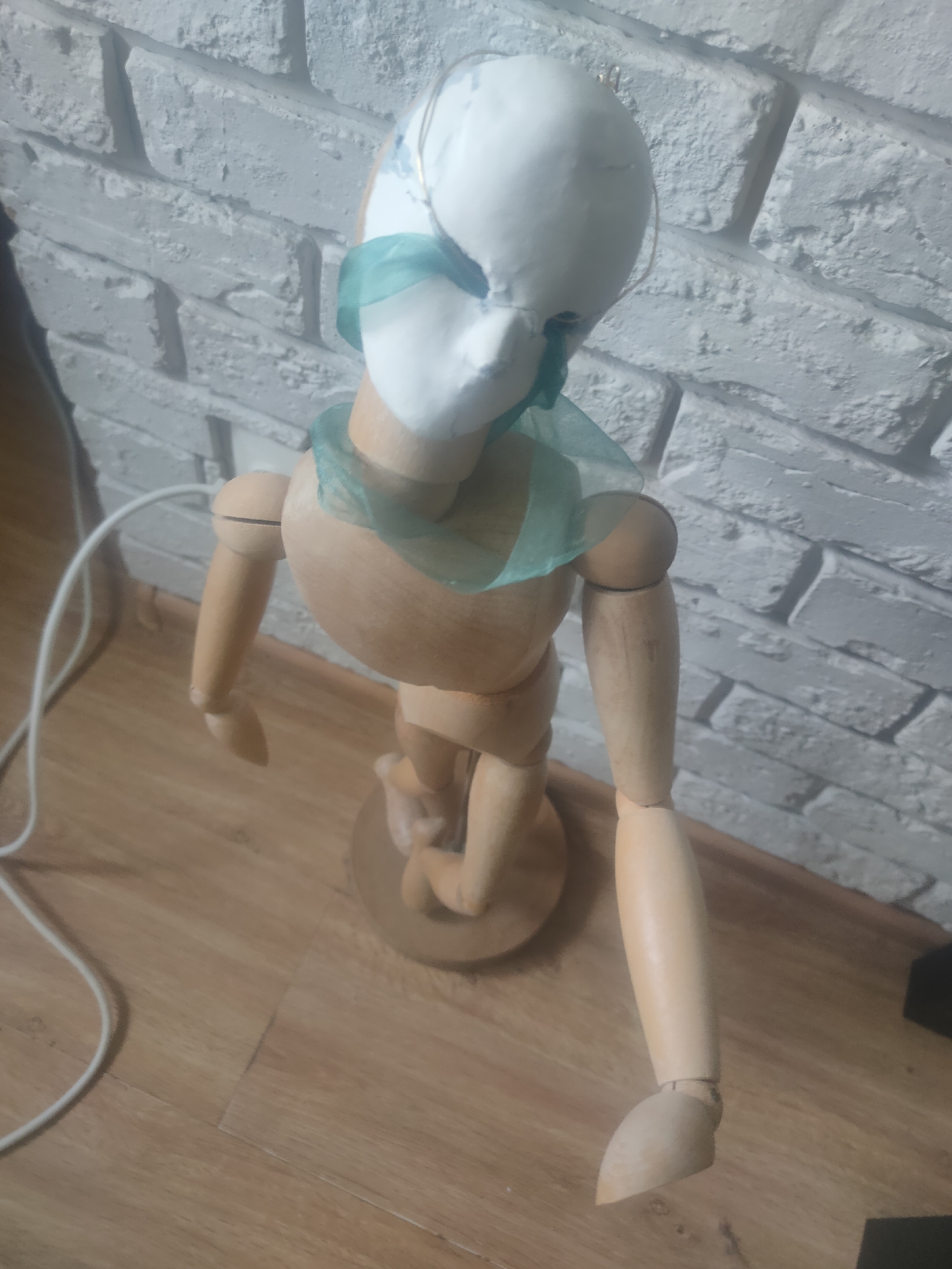 The image depicts a wooden mannequin with a white, mask-like covering on its head, adorned with a sheer, pale green fabric around its neck. The mannequin is standing on a wooden floor in front of a white brick-patterned wall. A white cable snakes across the floor to the left of the mannequin. The scene appears to be indoors, in a room that could be a studio or workshop. There is no indication of the city of Magadan, Russia, or any other specific geographical location.