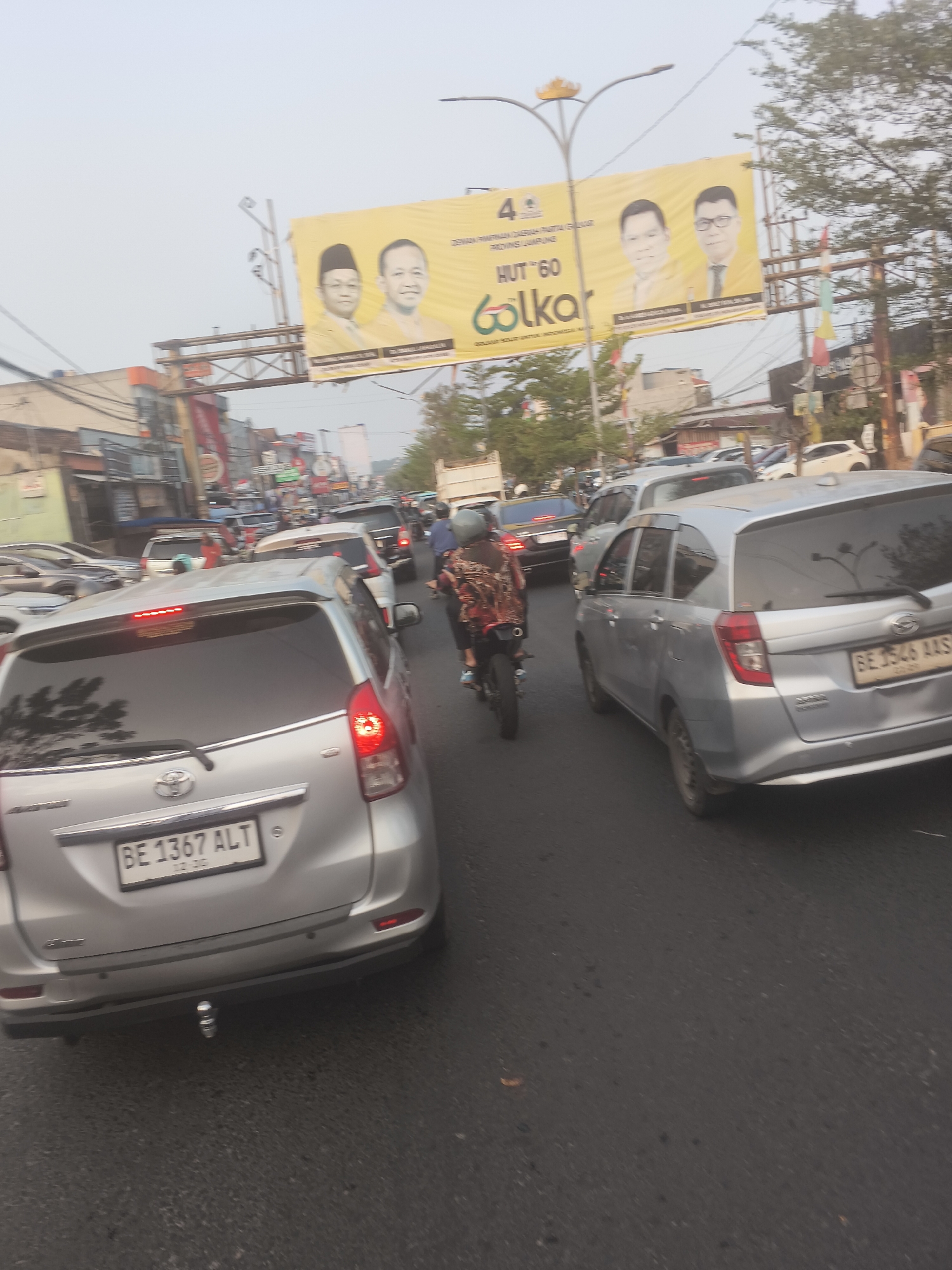 The image depicts a busy street scene in Bandar Lampung, Indonesia, likely during daylight hours given the overall brightness and shadows. The primary focus is on a traffic jam, with numerous vehicles including cars and motorcycles filling the road. The foreground features the rear of a silver Toyota Avanza with the license plate "BE 1367 ALT". To its right, another silver car with license plate "BE 1546 AAS" is visible. Further back, other vehicles and a motorcycle with a rider wearing a patterned shirt are present.
Dominating the upper portion of the image is a large yellow banner advertising "HUT '60 Golkar" and featuring portraits of two men. The banner is positioned above the traffic, suggesting it's part of a street-level advertisement or celebration. The environment appears to be an urban setting with buildings and trees lining the street, some of which are partially obscured by the vehicles and the banner. The sky is overcast or hazy, contributing to the diffuse lighting. The overall impression is one of congestion and daily urban life in Bandar Lampung.