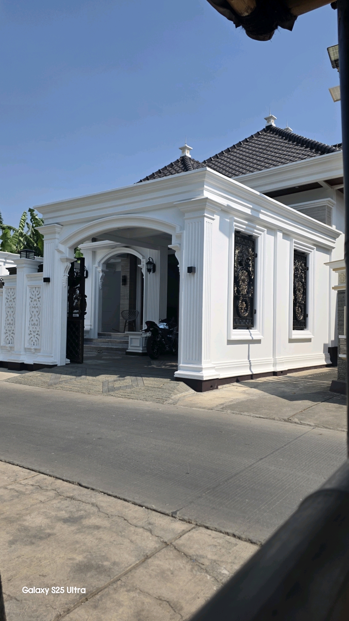 The image shows a modern, white house with a dark-tiled roof and ornate black metal window grilles. The house features a covered entrance with pillars and an arched opening. A black motorcycle is parked under the entrance. The property is bordered by a white, decorative fence. The foreground shows a paved driveway and a road. The sky is clear blue. The image also includes a watermark at the bottom left corner that reads "Galaxy S25 Ultra". This is a property located in Juwana, Indonesia.