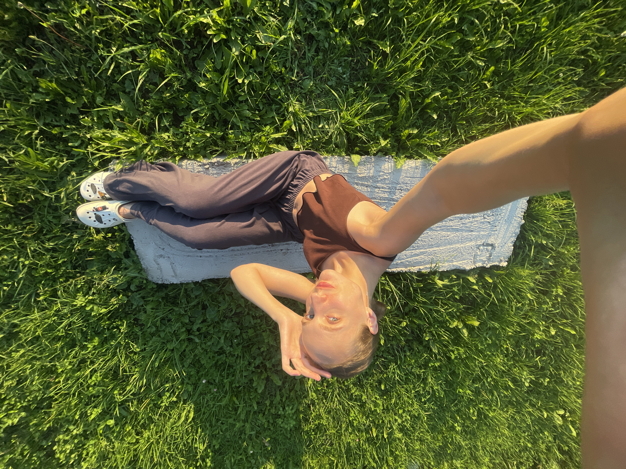 The image shows a young woman lying on a light-colored mat on a grassy area, outdoors. She is wearing a brown tank top and dark pants, with white slip-on shoes. Her head is resting on her hand, and her body is oriented vertically in the frame. The background is a dense green lawn, with patches of clover and other low-lying vegetation, bathed in warm sunlight. The overall setting appears to be a park or open green space.