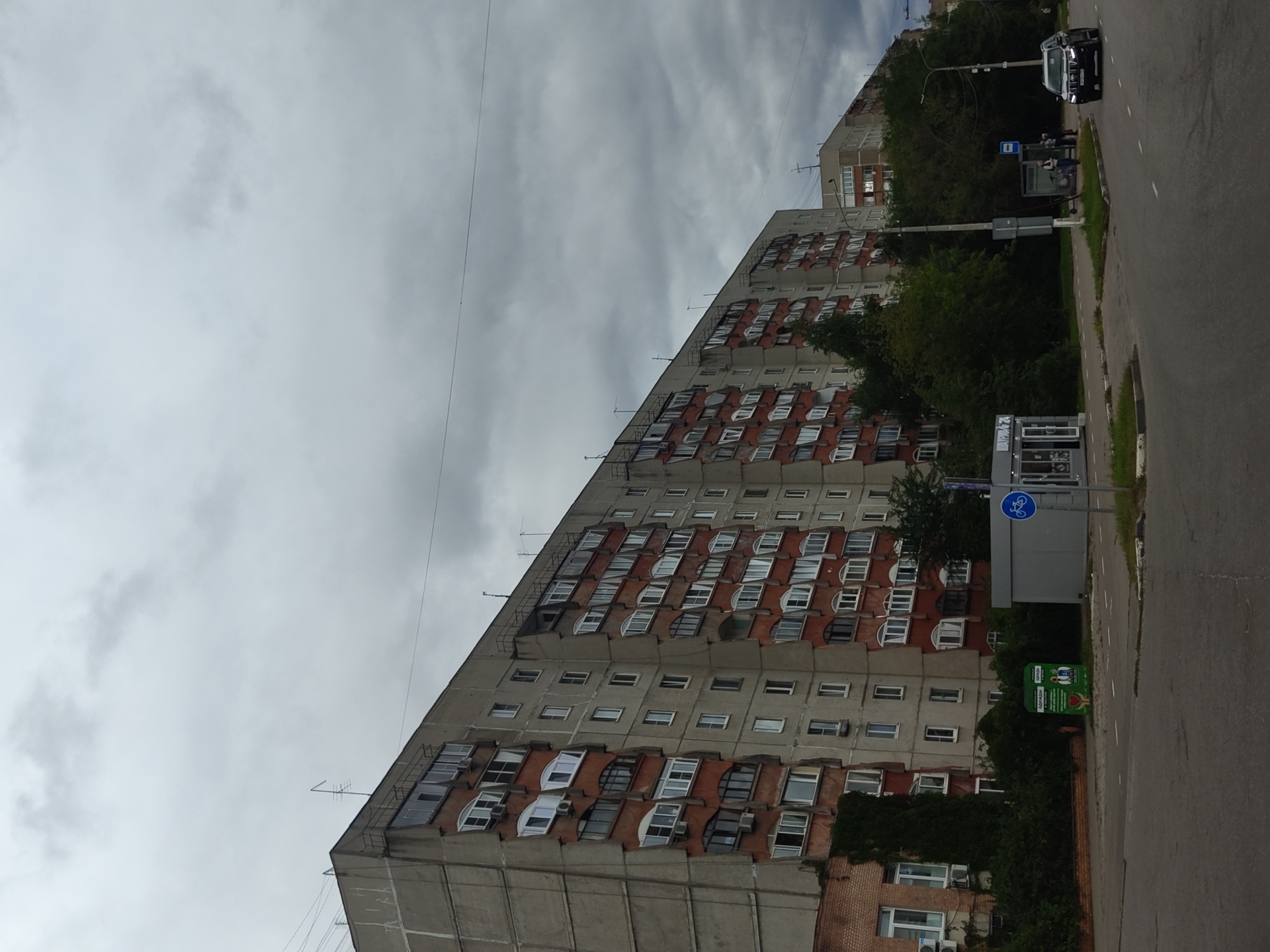A wide, overcast day scene in Zhukovsky, Russia, features a large, multi-story residential building dominating the background. The building's facade is characterized by a mix of grey concrete and red panels, with numerous windows and balconies. To the right of the residential building, a smaller, lighter-colored building is visible. In the foreground, a paved road with white markings runs across the image. A dark SUV is driving on the road. Adjacent to the road are sidewalks and green spaces with trees. A small kiosk with glass doors and a sign displaying a bicycle symbol is situated on the sidewalk. Further along the sidewalk, a bus stop with a shelter and a few people waiting can be observed. A green recycling bin with an image of people and a heart is also present on the side of the road. The sky is heavily clouded, suggesting a dull or rainy day.