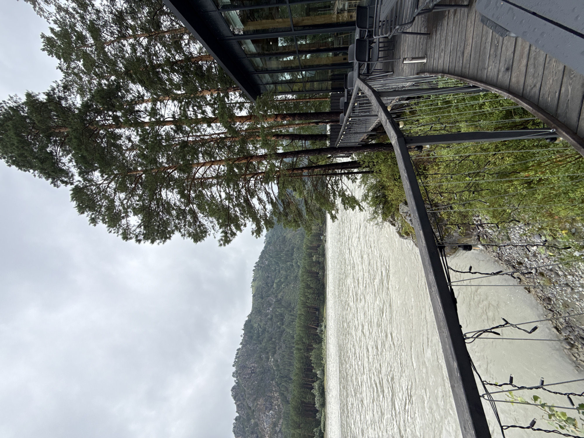 An outdoor seating area with tables and chairs is situated on a wooden deck next to a river. Tall pine trees line the edge of the deck, and a modern building with large glass windows is visible behind them. The river is wide and appears to be flowing rapidly, with a mountain covered in trees in the background. The sky is overcast. String lights are hung along the railing of the deck. The scene is likely in a natural, possibly mountainous, area of Russia, given the mention of Nizhnekayancha.