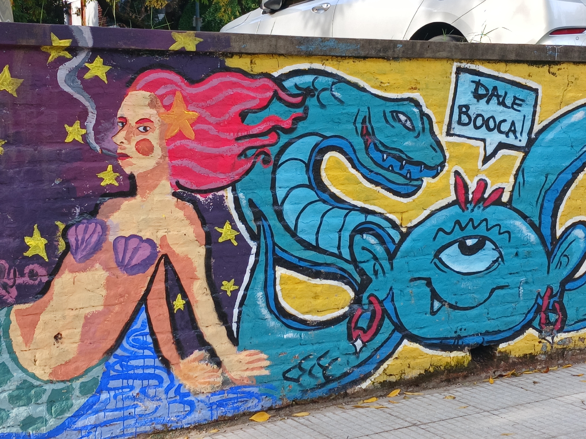 A vibrant mural adorns a brick wall in Buenos Aires, Argentina. On the left, a mermaid with red hair and a starfish in her hair is depicted smoking. Her skin is yellowish-orange, and she wears purple seashells. The background behind the mermaid is purple with yellow stars. To the right, a blue dragon-like creature coils, with an adjacent speech bubble reading "DALE BOOCA!" Below the dragon is another blue creature with one eye. The wall is yellow in this section. The mural is situated above a paved walkway and has a section of a white car visible above.