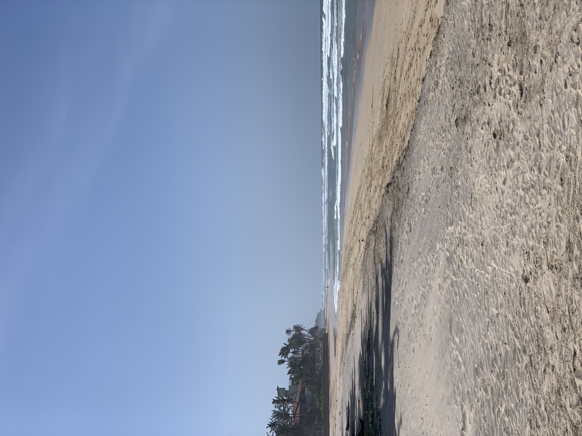 This is a vertical, eye-level, full shot of a wide, sandy beach in Kathaluwa, Sri Lanka, under a clear blue sky. The sand in the foreground is light brown and textured with ripples. Further back, shadows from unseen trees fall across the sand. Palm trees line the coast in the middle ground, with a building, possibly a hotel, visible among them. The ocean stretches to the horizon, with white-capped waves breaking on the shore. The water is a muted blue-green, and a hazy mist hangs over the distant sea. A single person is visible walking near the water's edge.