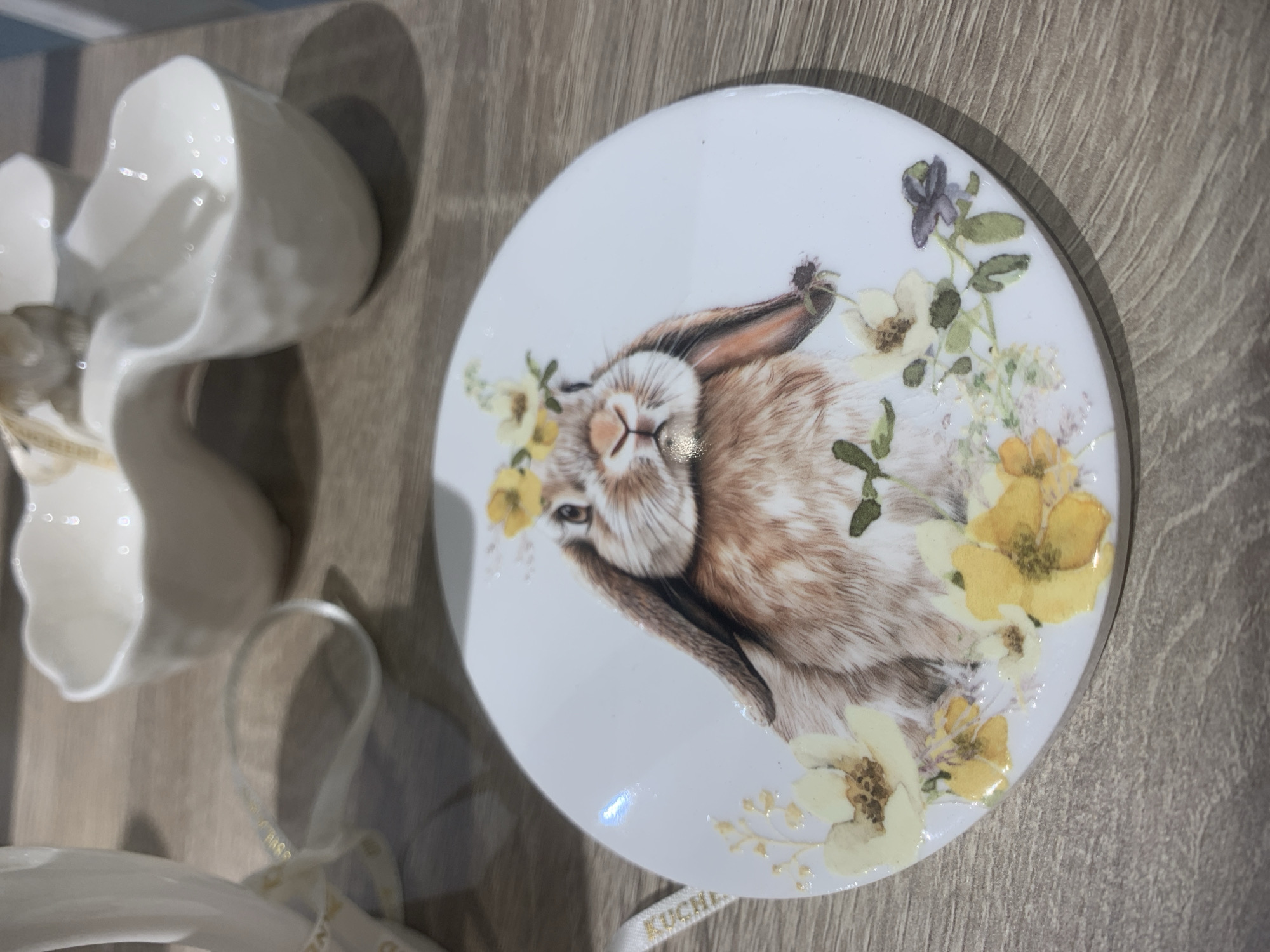 The media file displays an overhead, slightly angled view of decorative items arranged on a light brown, wood-textured surface. The photo was taken in Izhevsk, Russia. The primary focal point is a white, circular coaster or small plate, prominently featuring a detailed illustration of a light brown and white lop-eared bunny. The bunny's head is shown tilted, and its body is encircled by an artistic arrangement of flowers, predominantly in shades of yellow, cream, and faint purple, some appearing to have a glossy or raised texture.
To the left of the coaster, partially visible, are several white, reflective ceramic or porcelain cups/bowls, distinguished by their organic, wavy top edges. These items cast distinct shadows on the wooden surface. In the bottom foreground, a light-colored ribbon, possibly white or cream, is visible. Golden text printed on the ribbon clearly shows the word "KUCHE" alongside other partial letters. The lighting suggests an indoor setting, with bright reflections on the shiny surfaces of the objects. The overall composition suggests a theme of home decor, possibly for spring or Easter.