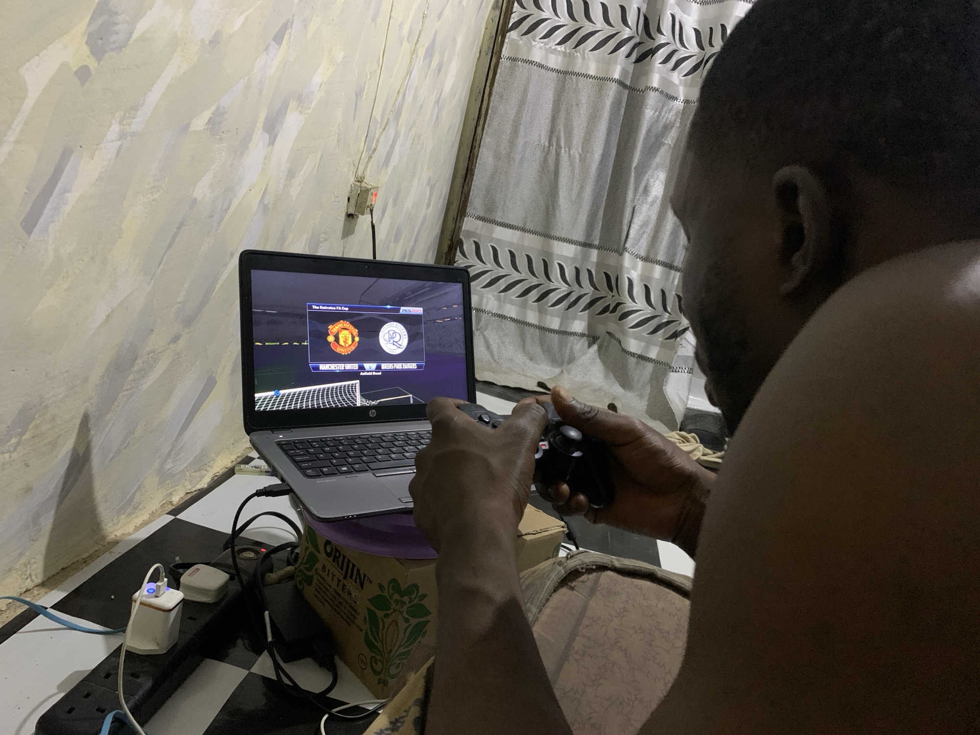 Here's a description of the image:
An African man is playing a soccer video game on a laptop. The screen displays a match between Manchester United and Queens Park Rangers. He is holding a black gaming controller. The laptop sits atop a cardboard box labeled "ORIGIN BITTERS." Cords and a charger surround the box and laptop, connected to a black power strip. The room has textured walls and striped curtains. The floor is covered in black and white checkered tiles. This is taking place in Rafin Par, Nigeria.