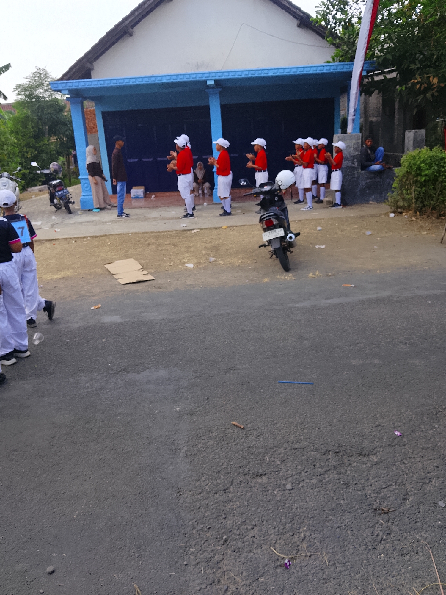 In Bangsri, Indonesia, a group of children in white caps and red shirts with white shorts stand in a line facing a building with blue trim and a dark garage door. Some of the children have their hands raised, palms open, as if in a performance or greeting. Behind them, an adult in a hijab stands next to a man in a dark shirt and blue jeans. A black motorcycle is parked to the left of the group, and another motorcycle is visible to the right, parked further back. The ground is covered in dirt and asphalt, with scattered debris including a cardboard box and some wrappers. A few adult onlookers are present further in the background.