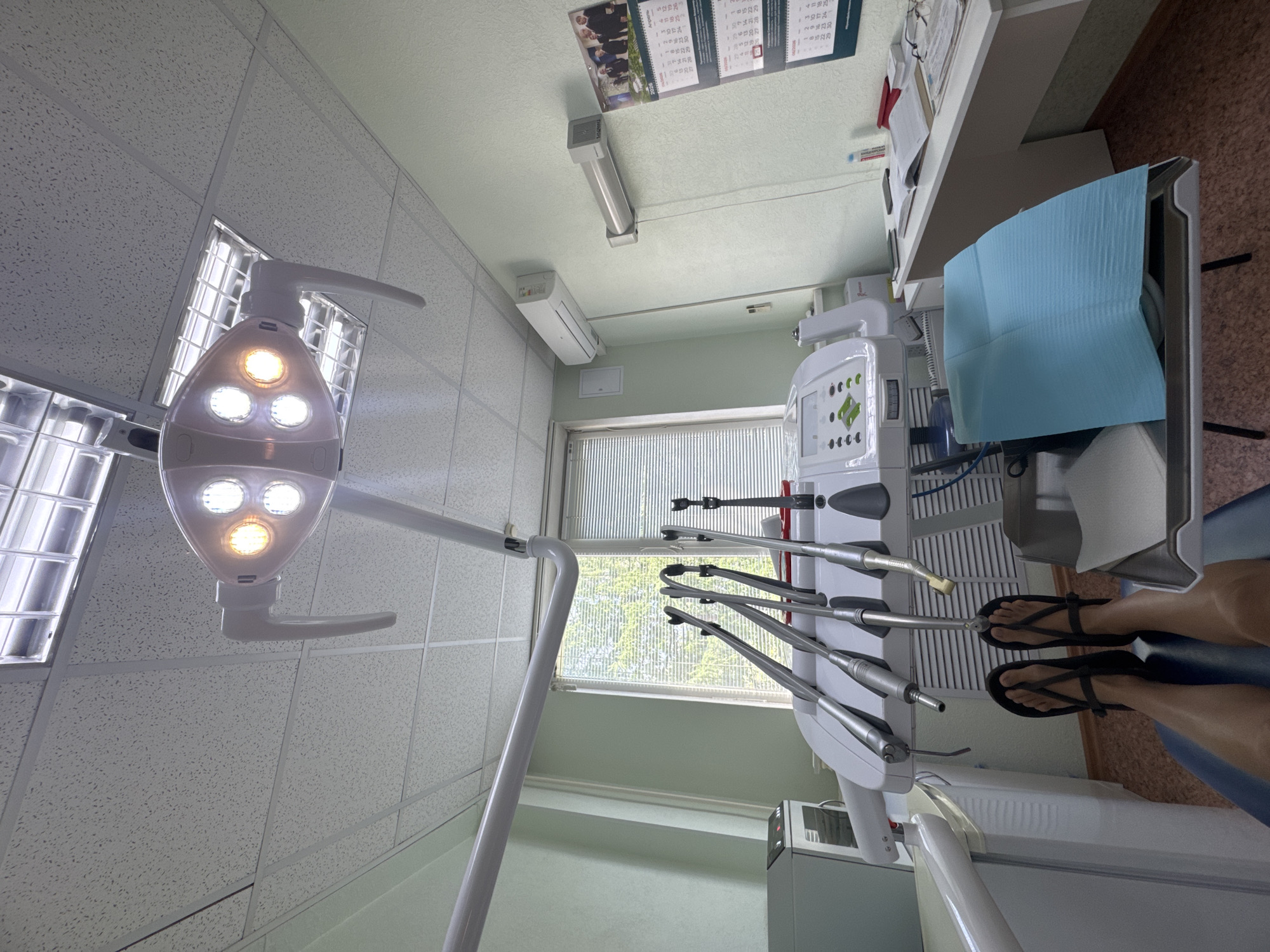 The image shows the interior of a dental office. A patient is seated in the dental chair, with their legs and feet visible in black sandals. Various dental instruments extend from the machine, positioned close to the patient. An examination light is focused on the patient. The room also contains office furniture like a desk and a window with blinds. A calendar hangs on the wall. The office appears clean and well-lit with drop ceiling tiles and fluorescent lighting. The location is in the city of Unknown, Unknown.