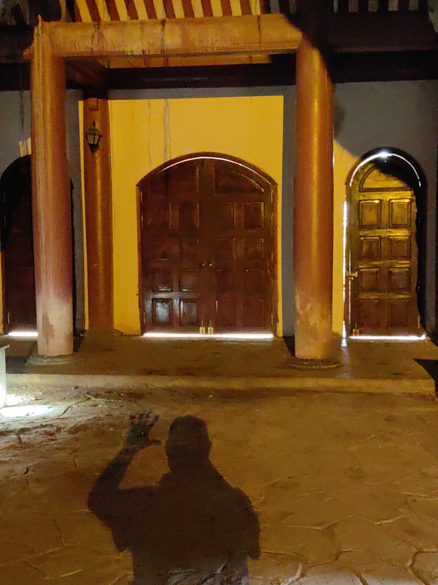 The image shows the entrance of a building in Lạng Sơn, Vietnam, at night. The building has three arched doorways. The central doorway has a large, double, wooden door. A light fixture is visible on the wall next to the central doorway. Two round, red pillars support the structure above the doorways. In the foreground, the shadow of a person waving is visible on the ground. The ground appears to be made of stone or concrete. The lighting is dim and yellowish.