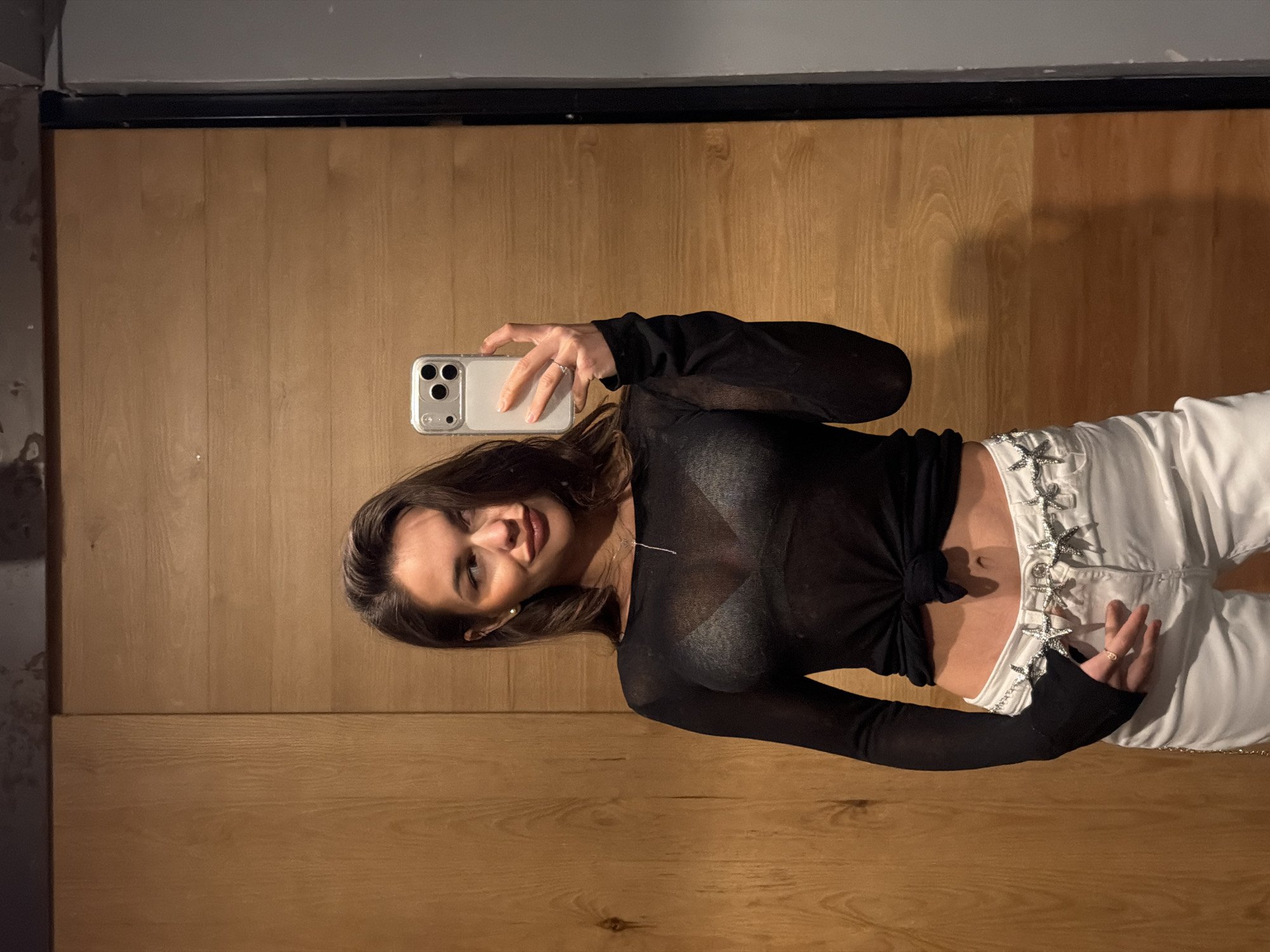 A woman takes a selfie in front of a wall with wood paneling. She is wearing a sheer black long-sleeved shirt tied at the waist, revealing part of her abdomen, and white pants with star-shaped decorations along the side. She holds a smartphone in her right hand. The location is not specified as Krasnoyarsk, Russia, and cannot be determined from the image.