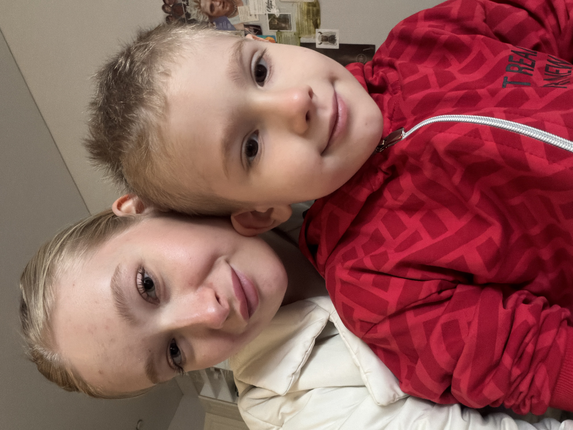 A young woman and a young boy are posing closely for a selfie indoors. The woman has blonde hair and is wearing a white puffy jacket. The boy has light brown hair and is wearing a red zippered jacket with a geometric pattern. They are both looking at the camera and smiling. The background is blurred, but appears to show a wall with photos or papers attached to it. Based on the model's knowledge, it is not possible to confirm whether the location is Засечное, Russia or any other specific location.