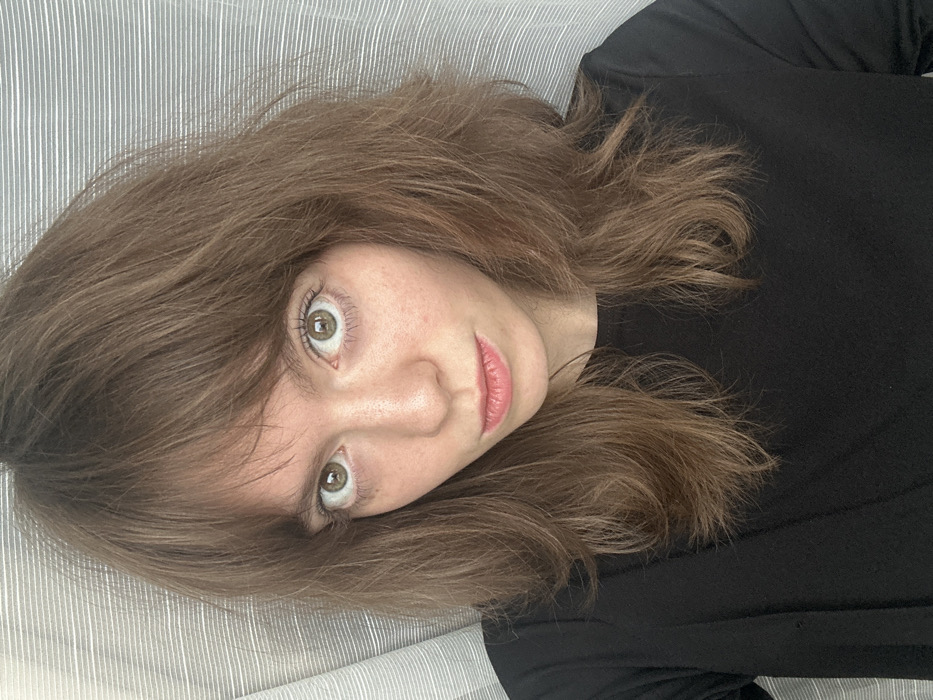 A young adult female is depicted in a frontal, head-and-shoulders portrait, looking directly at the viewer. She has light brown, shoulder-length hair with bangs, wide-set hazel eyes, and a fair complexion with visible freckles on her nose and cheeks. Her lips are slightly parted. She is wearing a plain black long-sleeved top. The background consists of a blurred, light grey fabric with subtle horizontal stripes, indicating an indoor setting. This scene is situated within the city of Moscow, Russia.