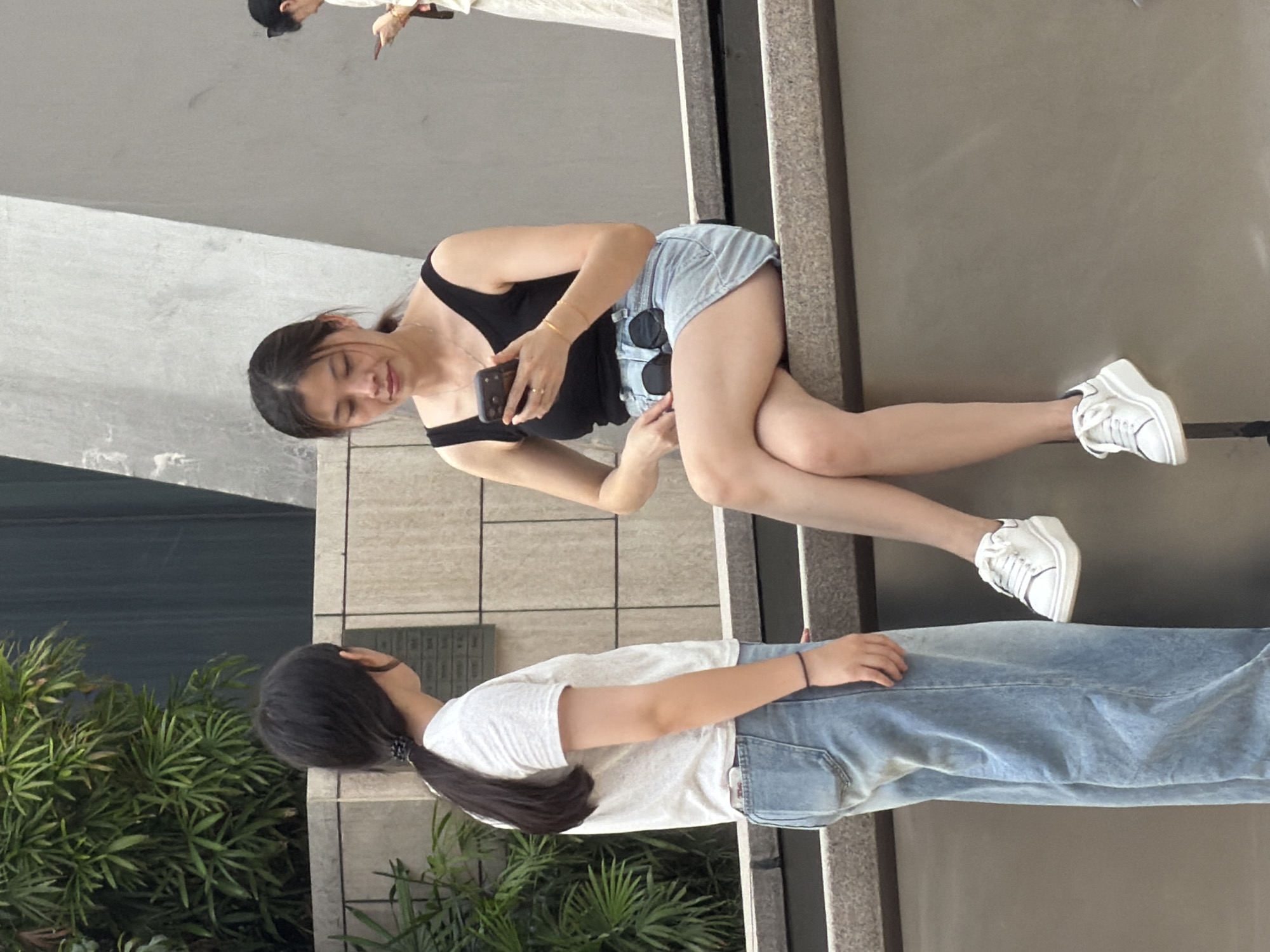 Two young women are interacting on an outdoor stone ledge. The woman on the left, standing, wears a white t-shirt and light blue wide-leg jeans. She has dark hair pulled into a ponytail. The woman on the right, seated, wears a black tank top and a light blue denim skirt. She has dark hair in a ponytail and is holding a smartphone. Her legs are crossed, and she is looking down at the phone. Behind the women are lush green foliage and a modern building with concrete walls. The scene appears to be in a public space in Yuzhong District, China.