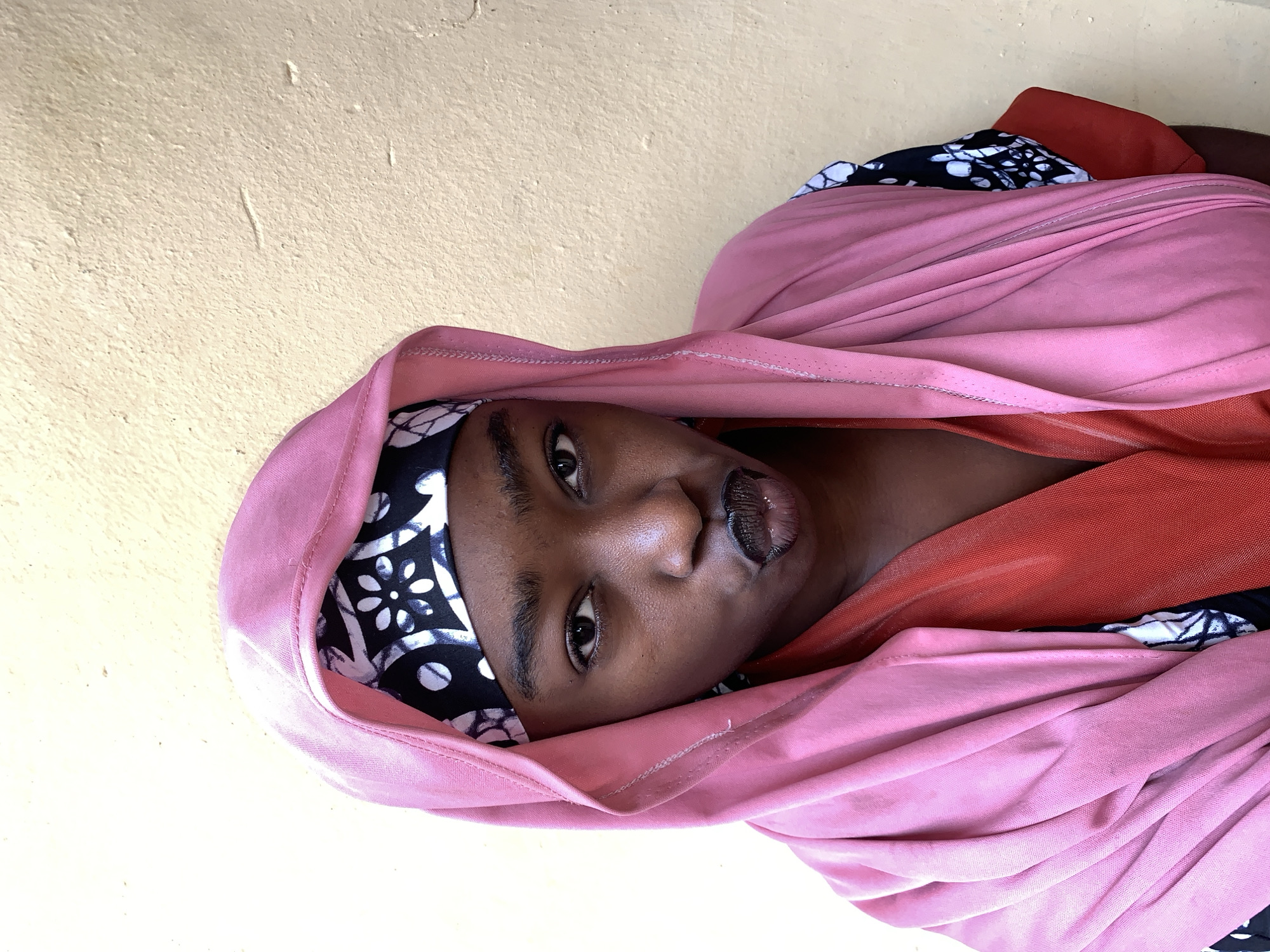 A medium shot shows a dark-skinned person wearing a pink hooded garment over a black and white floral headscarf. The garment frames their face. They have dark eyes and lips, and are looking slightly to the right. The person appears to be resting against a light yellow wall. The location and specific details of the environment within Rafin Par, Nigeria, cannot be determined from the given image.