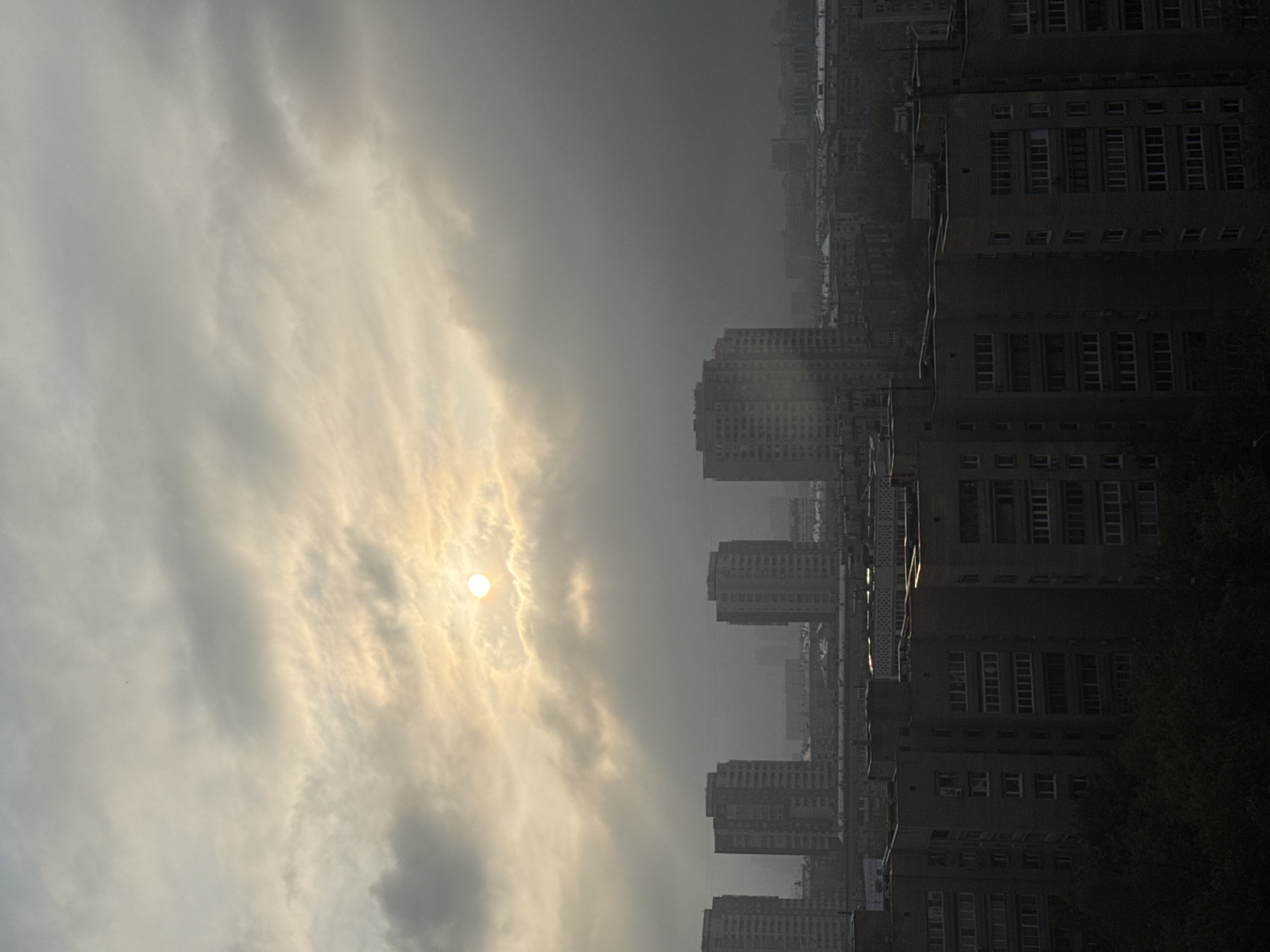 The image depicts a cityscape in Moscow, Russia, under a cloudy sky with the sun partially obscured by the clouds. Rows of high-rise apartment buildings dominate the right side of the image. The buildings are characterized by uniform, rectangular windows and a somewhat dated architectural style. The sky is filled with grey and white clouds, with the sun's light filtering through them, creating a hazy effect. The overall atmosphere suggests overcast conditions. No people are visible. The photograph is oriented sideways.