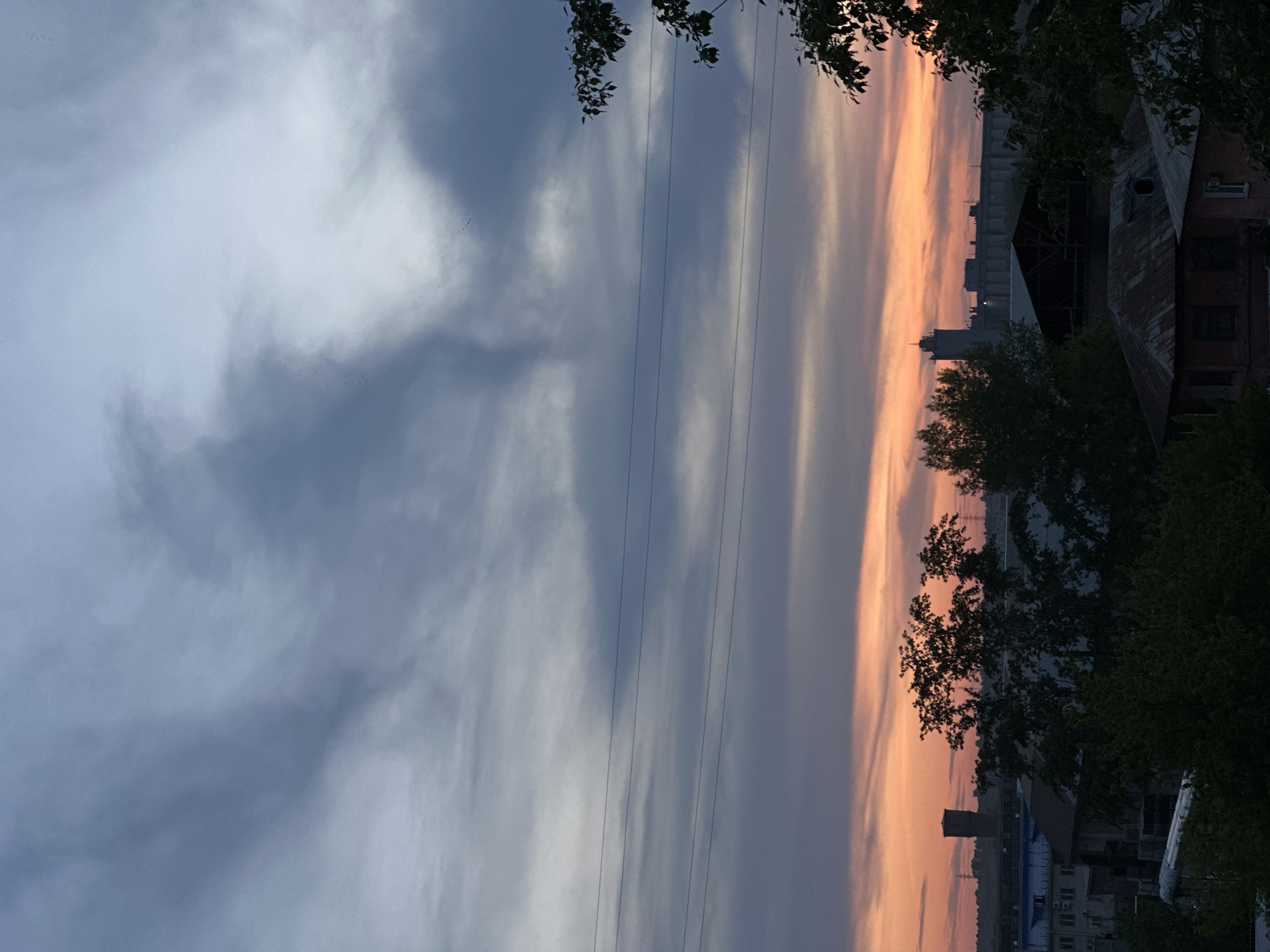 The image shows a twilight scene in Biysk, Russia. The sky transitions from a cloudy, grey-blue on the left to a vibrant orange-pink on the right, indicating sunset or sunrise. Power lines stretch across the sky from left to right. Silhouetted trees frame the upper and right sides of the image, partially obscuring buildings. A few buildings are visible, including a taller industrial structure with tanks and possibly residential buildings. The location is likely on the edge of town, where residential areas border industrial and agricultural land.