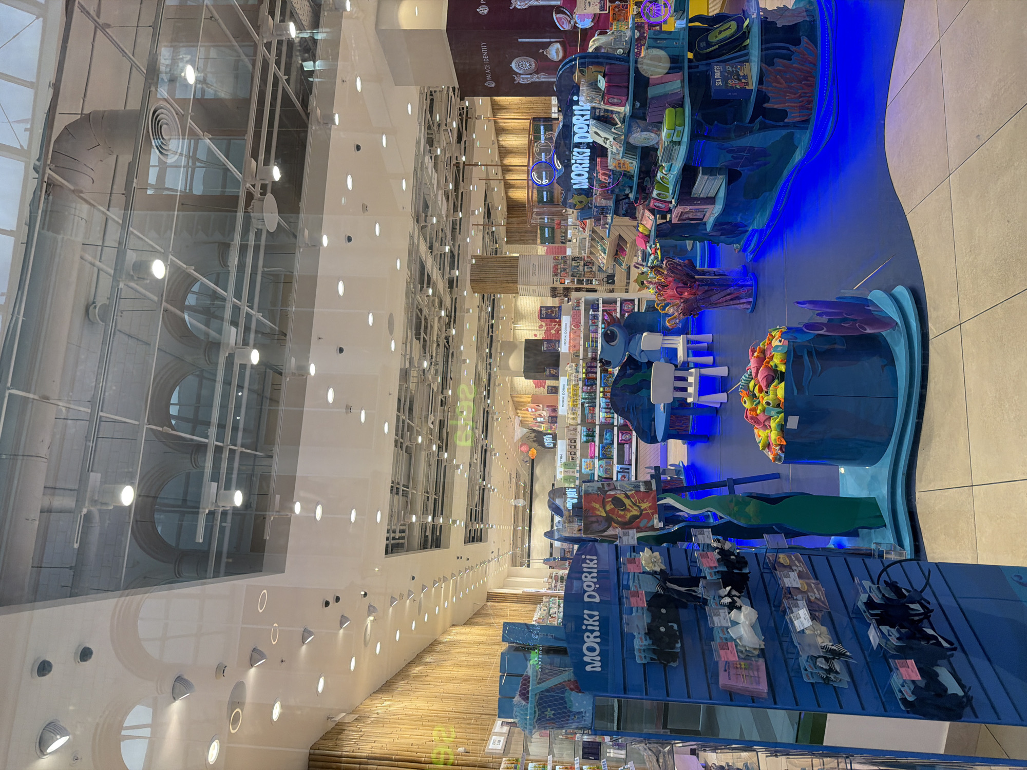 The image shows the interior of a brightly lit store, possibly a department store or a toy shop, with a focus on a display of children's items. The store features a high ceiling with exposed ductwork and lighting fixtures. The walls are decorated with a textured bamboo-like material and some shelves filled with merchandise. In the foreground, a blue display rack labeled "MORIKI DORIKI" holds various packaged hair accessories. Adjacent to this, a blue, wave-like display features children's toys, including plush sea creatures and a book titled "SEA PIRATES." Two small white chairs and a small blue table are also visible, suggesting a play or seating area. The floor is tiled with a light gray and beige pattern. The overall impression is of a vibrant and child-friendly retail environment.