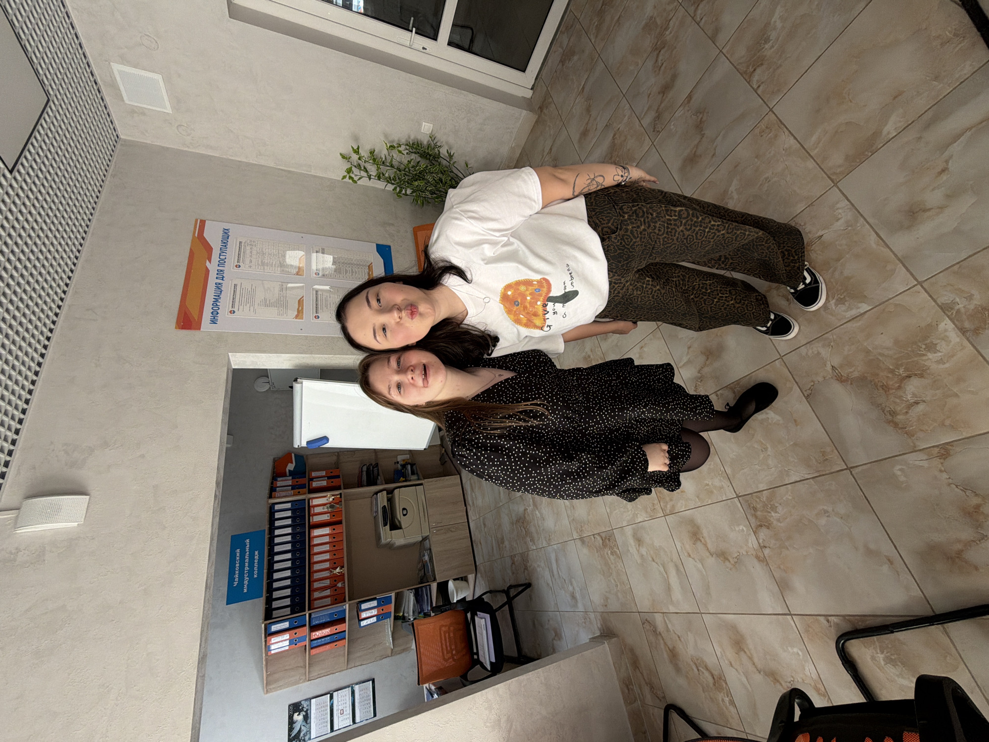 In an indoor setting, two young women are positioned in what appears to be an office or administrative area. The woman on the left, with light brown hair, wears a black dress with white polka dots and black tights and shoes. She is sticking her tongue out slightly. The woman on the right, with dark hair, wears a white t-shirt with a graphic of a mushroom and the text "give you a mushroom umbrella" and patterned trousers. She is puckering her lips.
Behind them, shelves are filled with binders and office supplies. A printer is visible on one of the shelves. A calendar hangs on the wall to the left, and a whiteboard is present to the right. A poster with the heading "ИНФОРМАЦИЯ ДЛЯ ПОСТУПАЮЩИХ" (Information for Applicants) is mounted on the wall. The floor is tiled with a beige and brown marble pattern. The overall environment suggests an institutional or educational facility, likely related to the "Чайковский индустриальный колледж" (Chaikovsky Industrial College) mentioned on a blue sign above the shelves. The lighting is bright and even, indicating an indoor environment with overhead illumination.