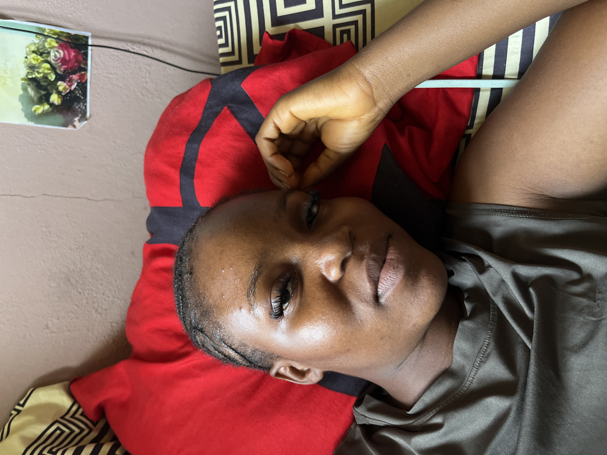 The image shows a close-up, overhead view of a young Black woman lying on a bed. She is looking towards the camera with a relaxed expression. She has braided hair and is wearing false eyelashes. Her right hand is raised and touching her temple. She is wearing a dark olive green t-shirt. Behind her, a red pillow with black accents is visible, as is a patterned pillow in shades of black and beige. A framed picture of flowers is attached to a light brown wall in the background. The scene appears to be indoors, likely a bedroom. The image does not provide any information about the location in Rafin Par, Nigeria.