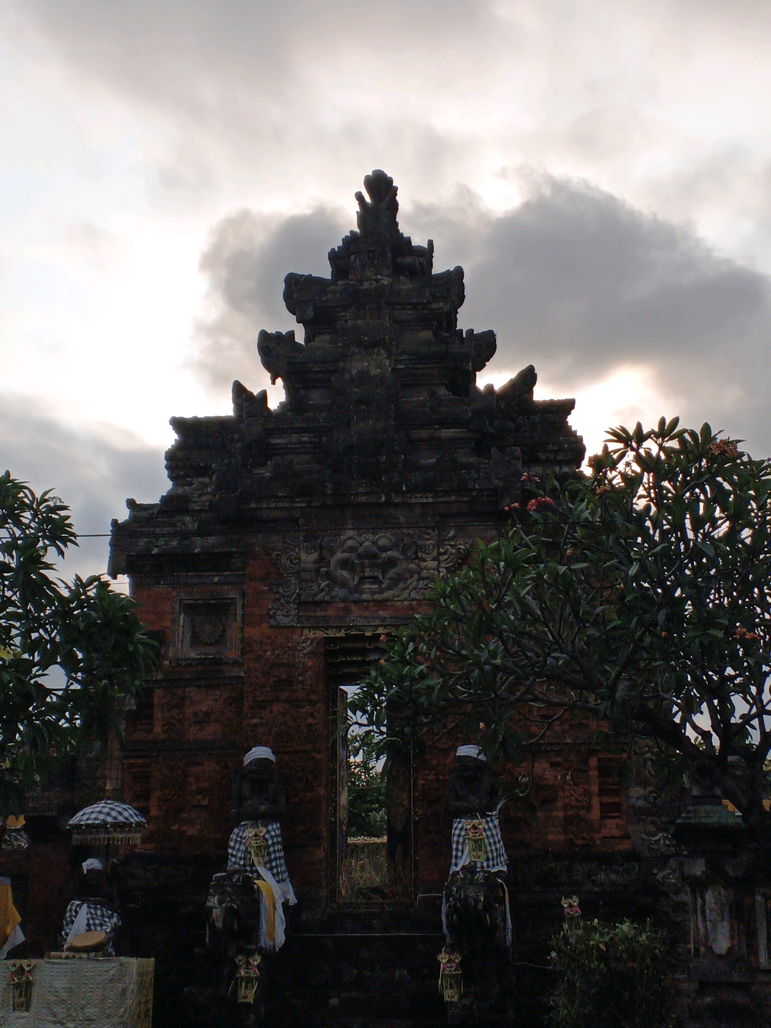 An outdoor, medium shot captures a Balinese temple gate in Blahbatuh, Indonesia, under a cloudy sky. The temple, constructed from reddish-brown brick and dark stone, features an intricate, multi-tiered roof adorned with carvings. Two weathered, statue-like figures stand guard at the base of the gate, each draped in traditional checkered sarongs and white head coverings. One figure is positioned to the left of the gate, near a checkered umbrella, and the other is to the right. In front of the statues, offerings or decorations are visible. Lush green foliage and flowering trees partially obscure the temple structure on the left and right sides of the frame. The overall lighting suggests either dawn or dusk, with the sun partially visible through a gap in the clouds, casting a soft glow.