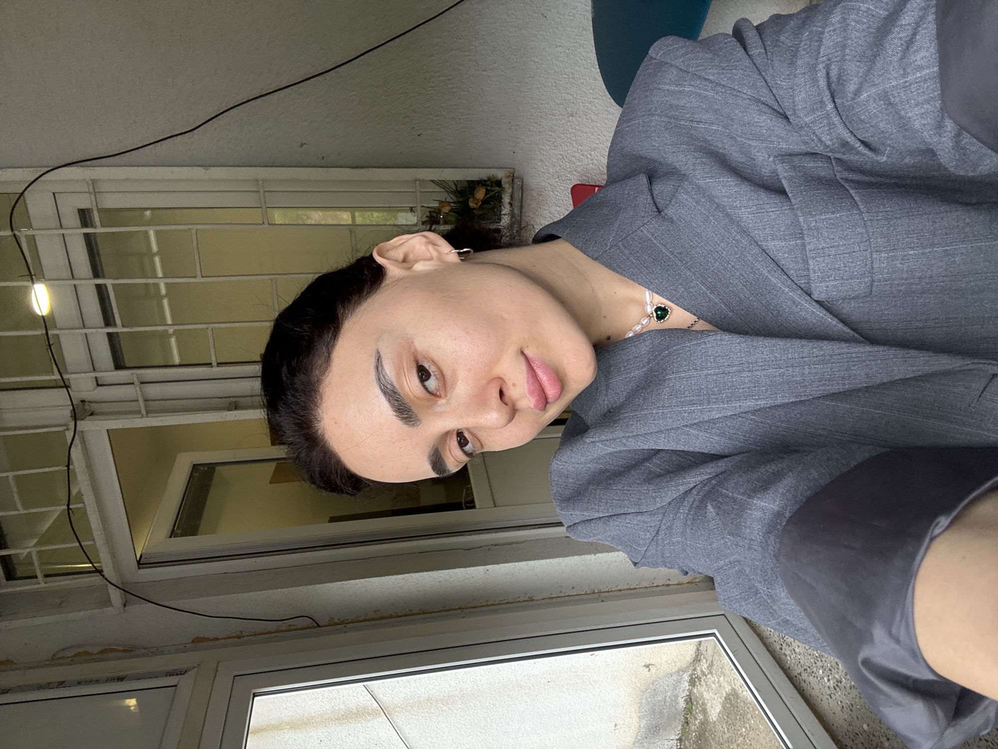 A person wearing a gray blazer and pearl necklace with a green pendant is indoors, possibly near a window. The window has a white frame and a grid pattern. The setting may be in Tbilisi, Georgia, but this cannot be confirmed without additional context clues. An electrical wire runs near the window. The person's expression is neutral. The details about the person's actions and interactions are not discernable from this image. The environment includes the interior space visible through the window and the exterior seen outside the window. The quality of the window frame suggests an older building.