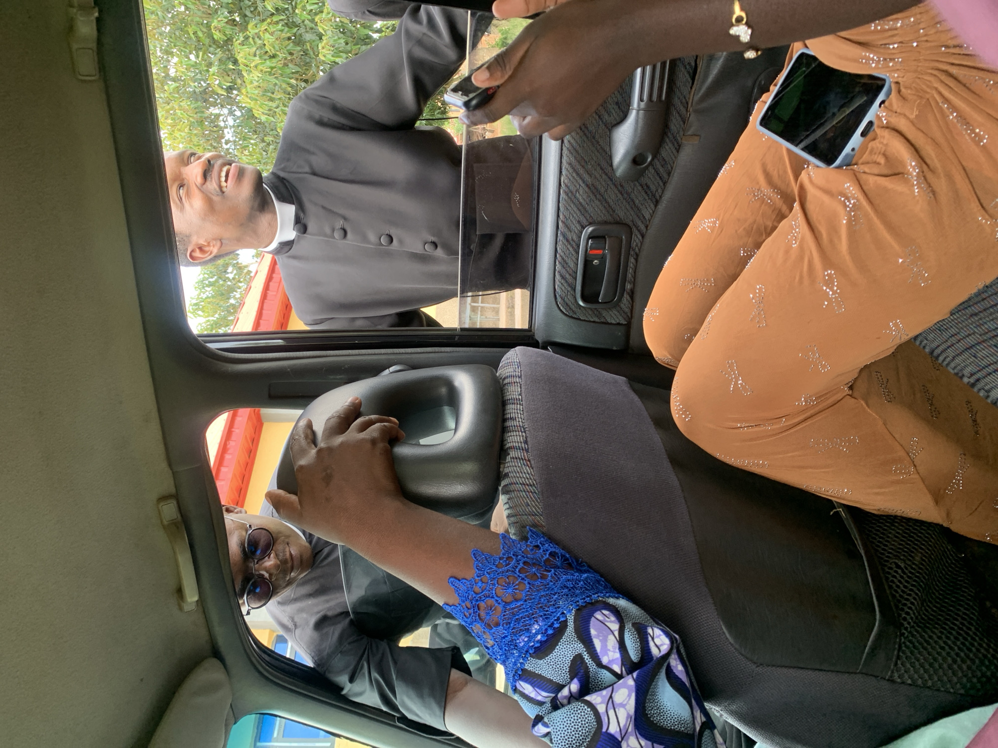 The image depicts an interior view of a vehicle in an outdoor setting. Two men, dressed in clerical attire, are visible through the car windows. The man on the left, wearing sunglasses, leans in towards the inside of the car, with his arm resting on the back of a car seat. The man on the right, smiling and looking away, is also positioned by the window. In the foreground, from inside the car, a person's legs are visible, wearing brown patterned pants, and a hand holding a smartphone. Another hand, adorned with a bracelet, is also seen. The environment outside the car appears to be a street with trees and buildings in the background.