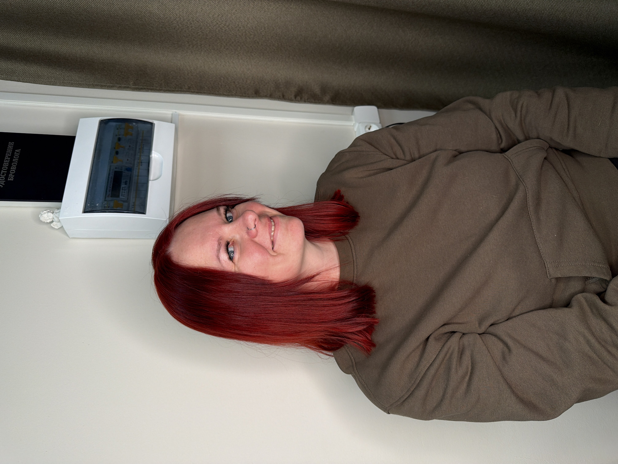 The image displays a woman situated indoors, likely within a beauty or cosmetology studio in Chaikovsky, Russia. The subject is a woman with bright red, shoulder-length hair, blue eyes, and a silver hoop nose piercing in her left nostril. She is wearing a plain olive-green crew-neck sweatshirt with a kangaroo pocket and is depicted smiling faintly while looking towards the viewer. The background features a light beige wall on the left and a dark olive-green curtain on the right. Mounted on the wall is a white electrical breaker box with a transparent cover, revealing internal components. Directly above the breaker box, a black folder or book titled "УДОСТОВЕРЕНИЕ БРОВ0ЛОГА" (BROWOLOGIST CERTIFICATE) is visible. A small white angel figurine is positioned to the left of the breaker box. A white electrical outlet with two sockets is also visible on the wall near the woman's right shoulder.