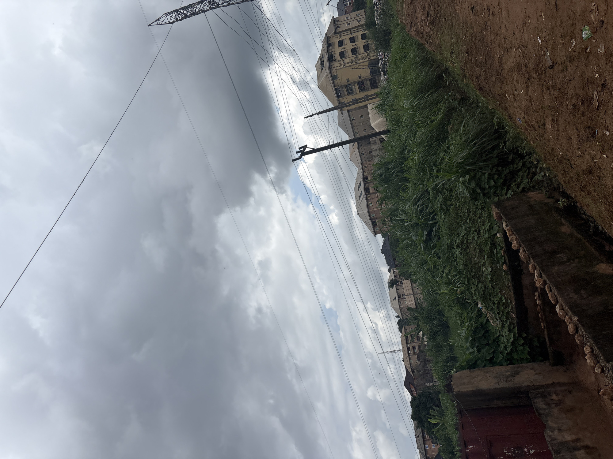 This is a tilted, eye-level outdoor shot of a cloudy sky over a residential area in Nkpor, Nigeria. The image is dominated by a vast, overcast sky, with thick, gray clouds filling most of the frame. In the mid-ground, several multi-story buildings with light-colored facades are visible, featuring numerous windows. Power lines crisscross the scene, supported by wooden poles. To the right of the buildings, lush green vegetation grows, bordering a dirt path. The path itself is reddish-brown and appears uneven, with scattered debris. A concrete structure with a reddish-brown door is visible in the foreground on the right. The overall impression is one of an overcast day in a developed, yet somewhat unkempt, urban or semi-urban environment. The time of day appears to be daytime, but the cloudy weather obscures direct sunlight. No specific activity or event is clearly depicted; the scene appears to be a static view of the surroundings. There is no visible text in the image.